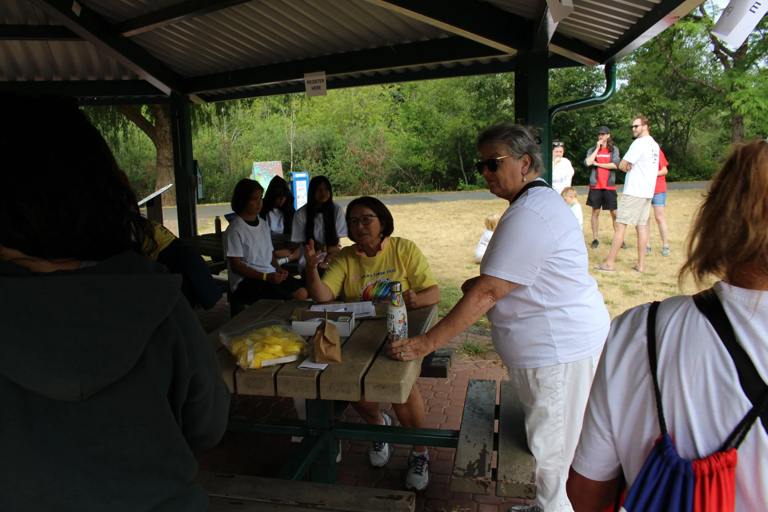 A group of people under a pavilion at an outdoor event, with some seated at a table and others standing or walking nearby, with trees and grass in the background.