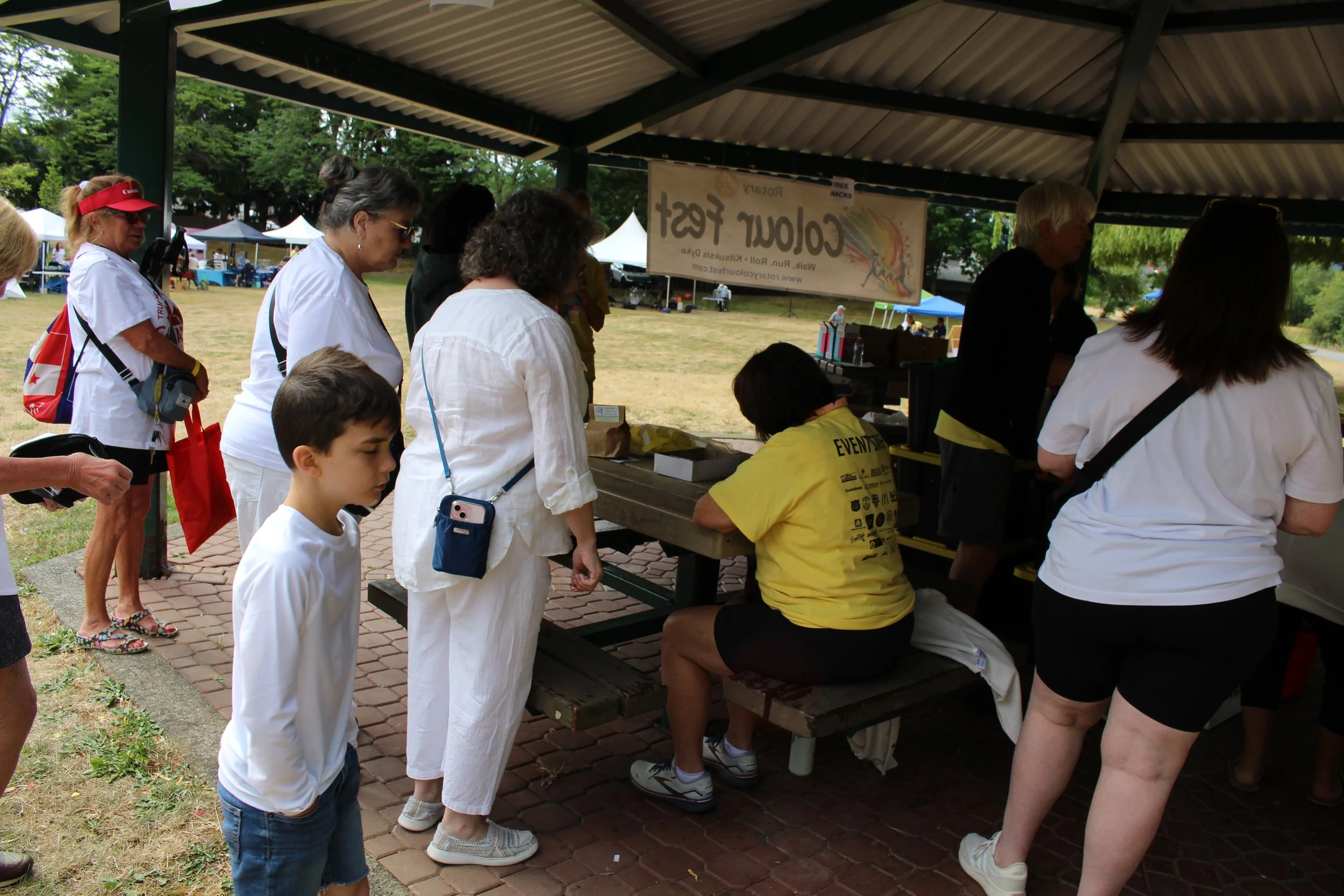 People standing in line at an outdoor event under a canopy. They are waiting at a booth where a woman in a yellow shirt is seated, possibly registering or purchasing something. There are trees and open fields in the background, with tents set up in t