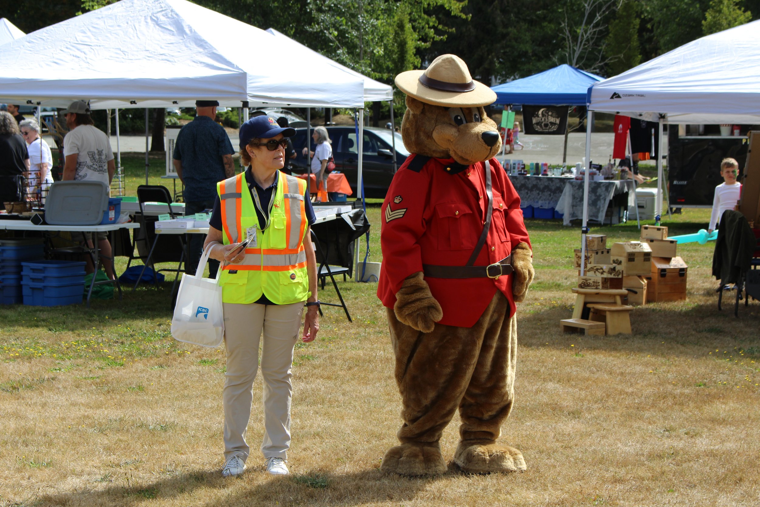 A woman in a yellow safety vest and khaki pants stands next to a person in a bear mascot costume dressed as a Canadian Mountie, outdoors during a sunny day at a fair or market with tents and other visitors in the background.