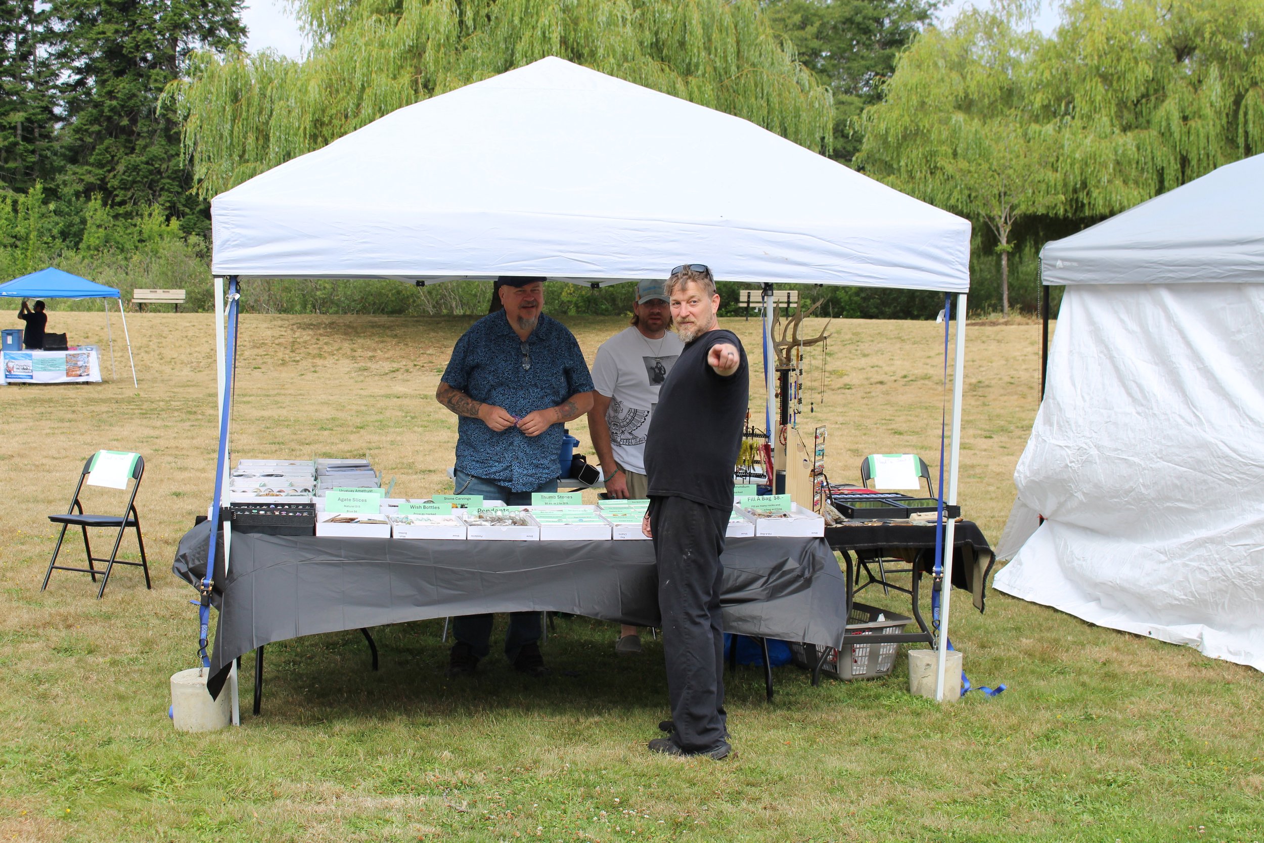 Man in black pointing at camera at outdoor market stall with three other men behind table selling jewelry and crafts, with tents and grassy field in background.