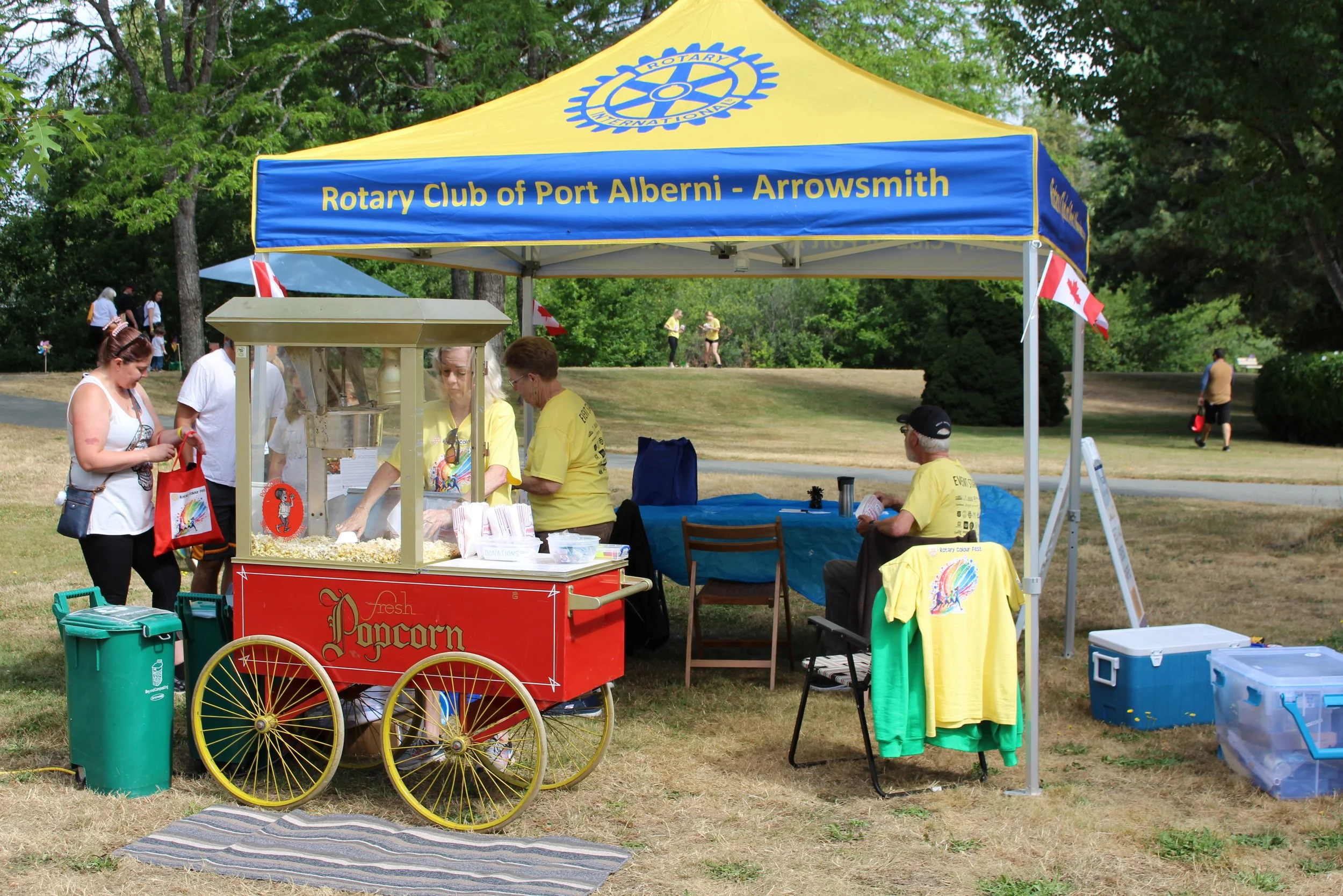 A popcorn stand at a Rotary Club event under a yellow and blue canopy that reads 'Rotary Club of Port Alberni - Arrowsmith'. Several people are gathered around, some serving popcorn and others sitting nearby. The scene is outdoors on a grassy area wi