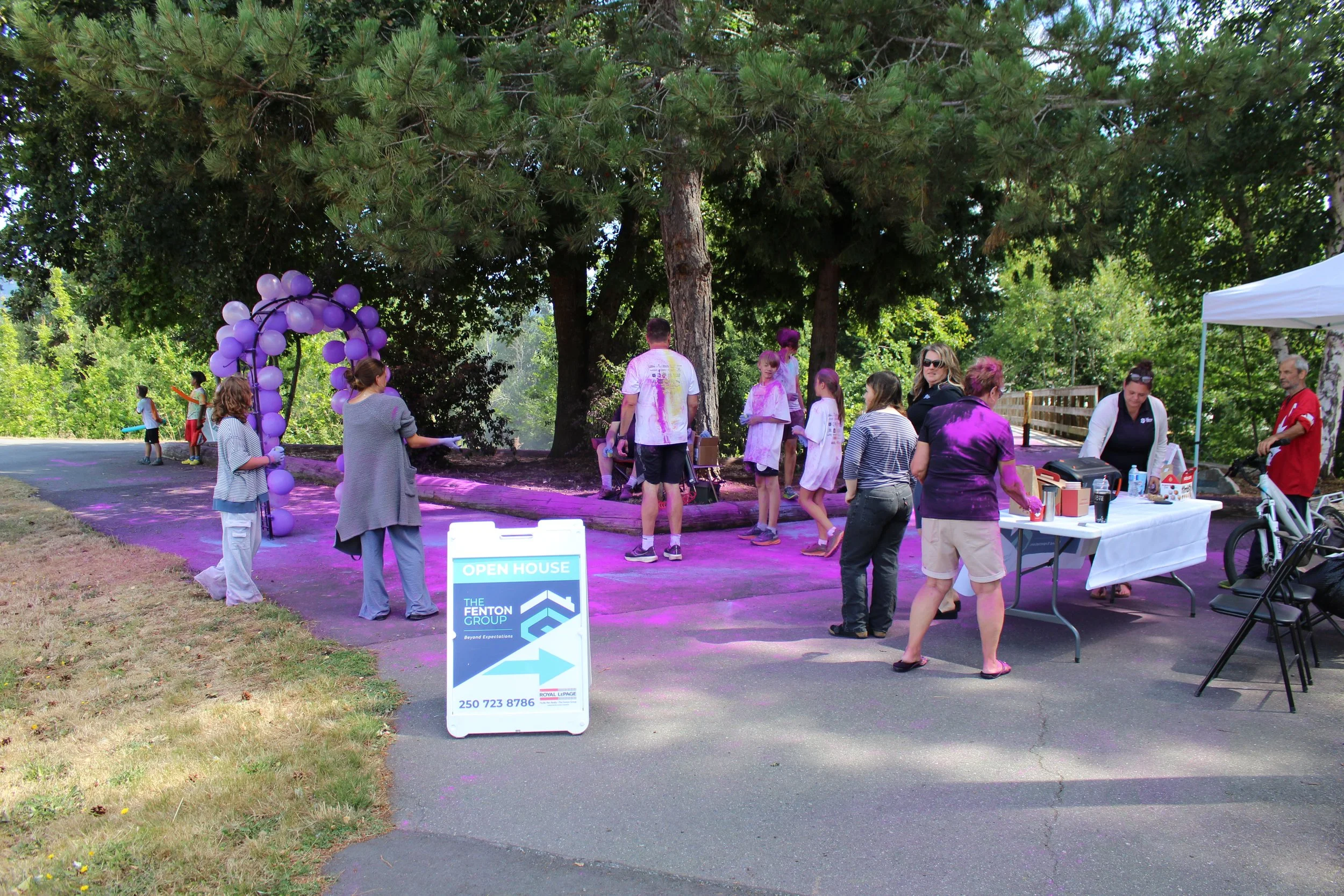 People gathered outdoors under trees for a color run event, with purple powder on participants and a balloon arch, as volunteers serve drinks at a table. A sign indicates an open house for The Fenton Group.