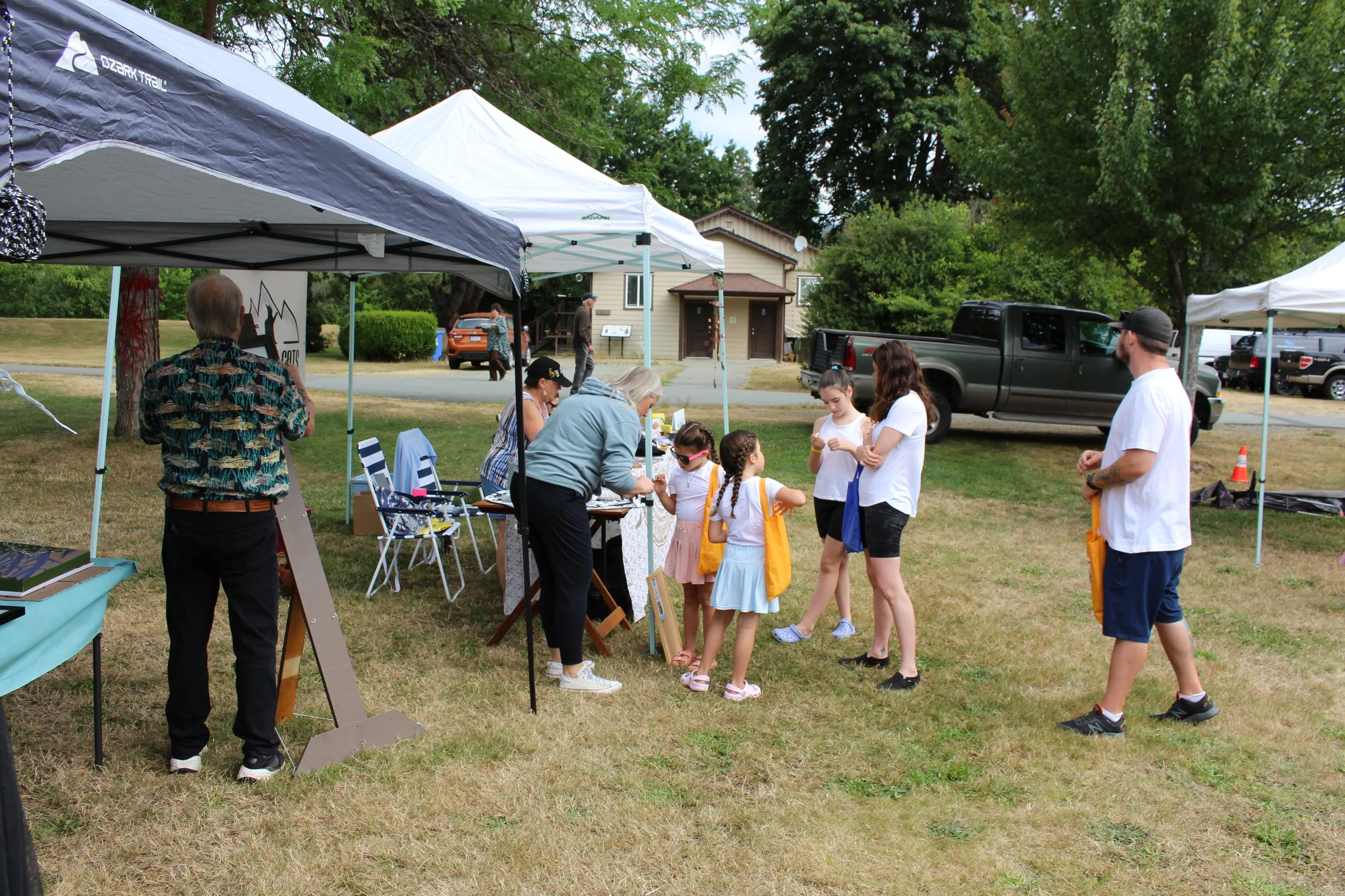People gathered at an outdoor community event with tents, tables, and children engaging in activities on a grassy area.
