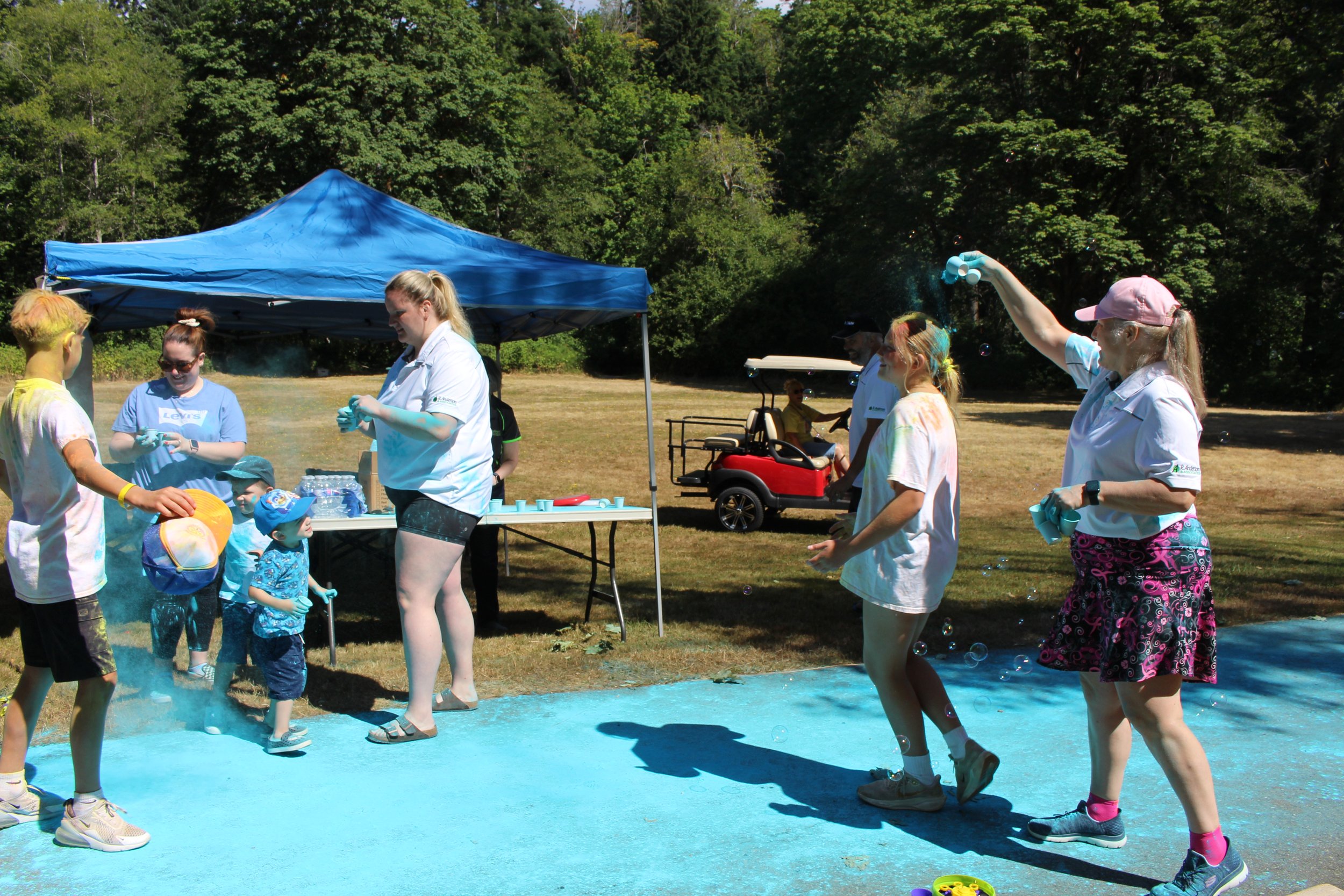 People participating in a color run event outdoors, throwing colored powder and bubbles, with a grassy field and trees in the background, and a blue canopy tent.
