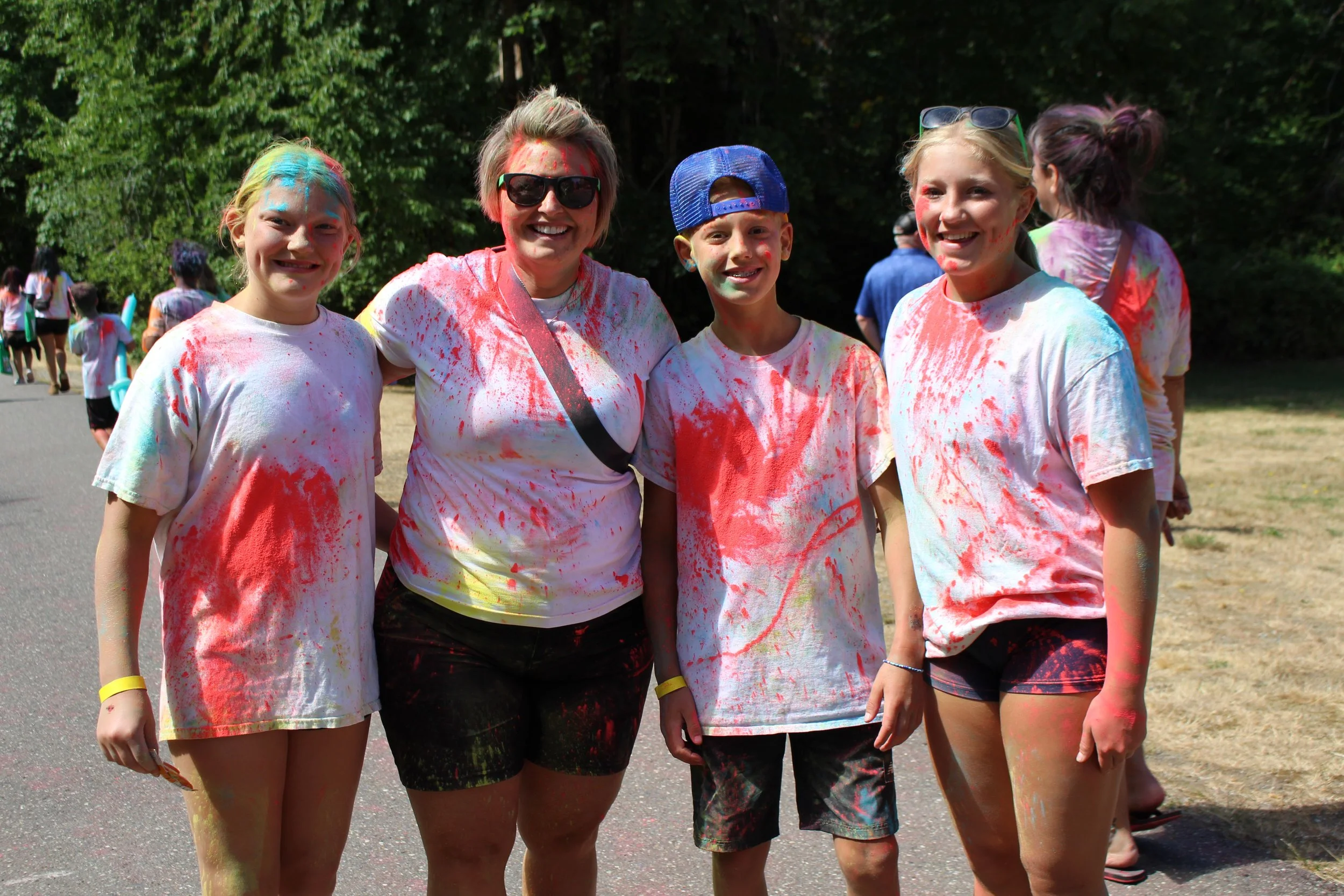 Group of five people, four young girls and one adult woman, smiling and covered in colorful powder at an outdoor event.