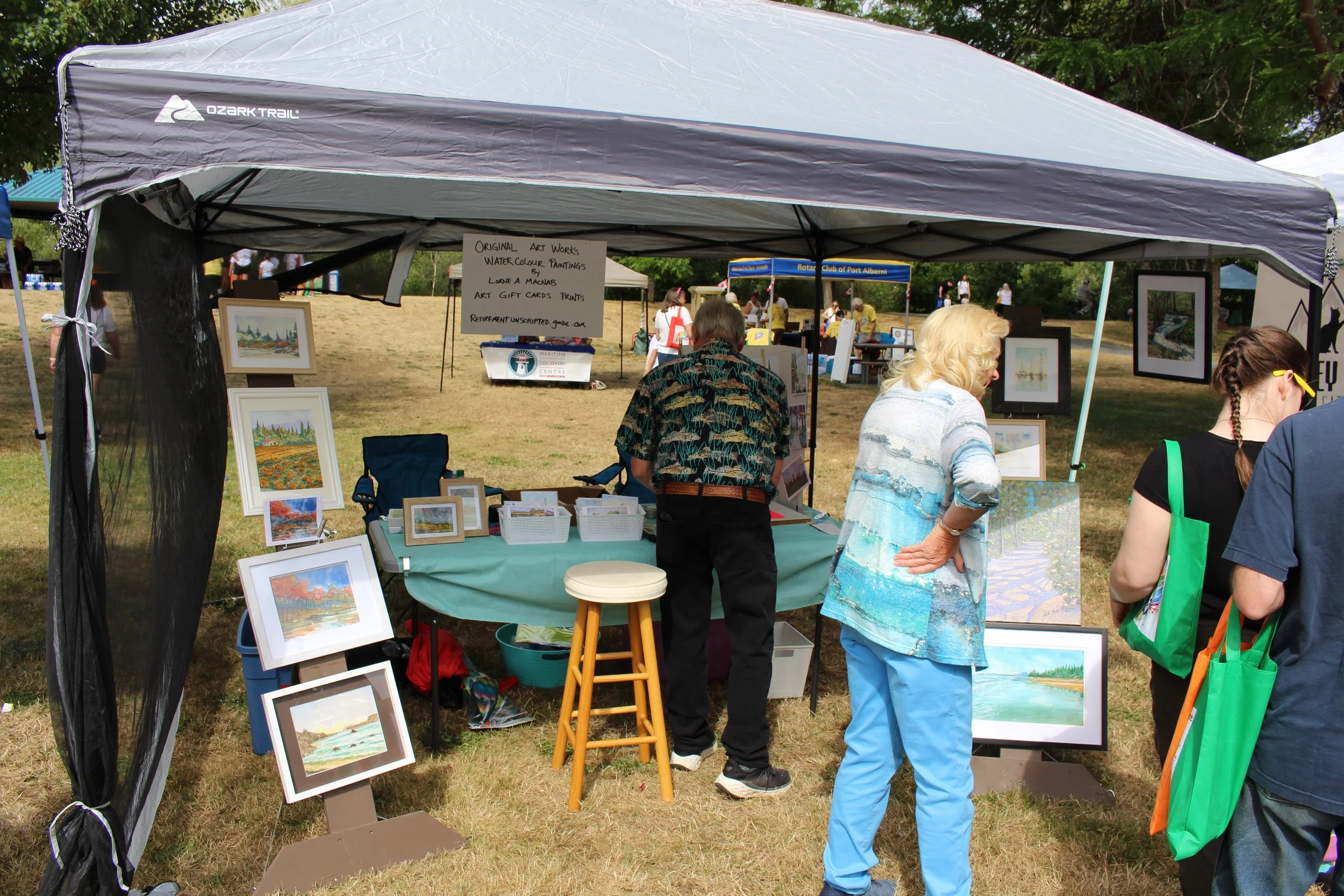 Art booth at an outdoor event with framed landscape paintings, a table with art supplies, and people viewing and discussing the artwork.