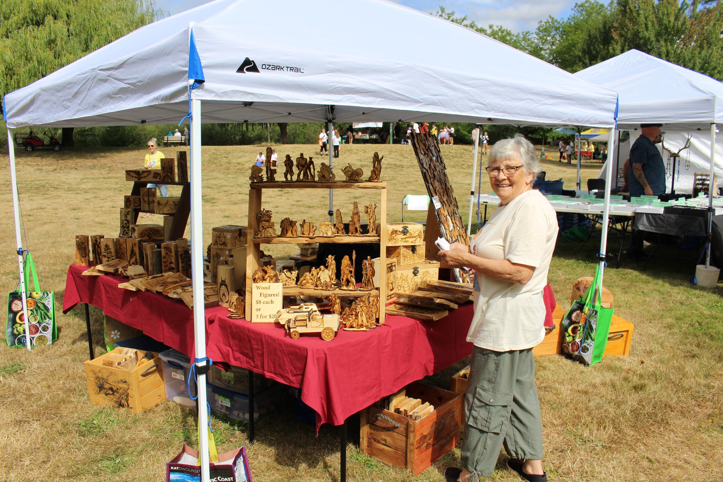 An elderly woman at an outdoor craft market stall selling wood-carved figurines and artwork under a white canopy, with other stalls and people in the background.