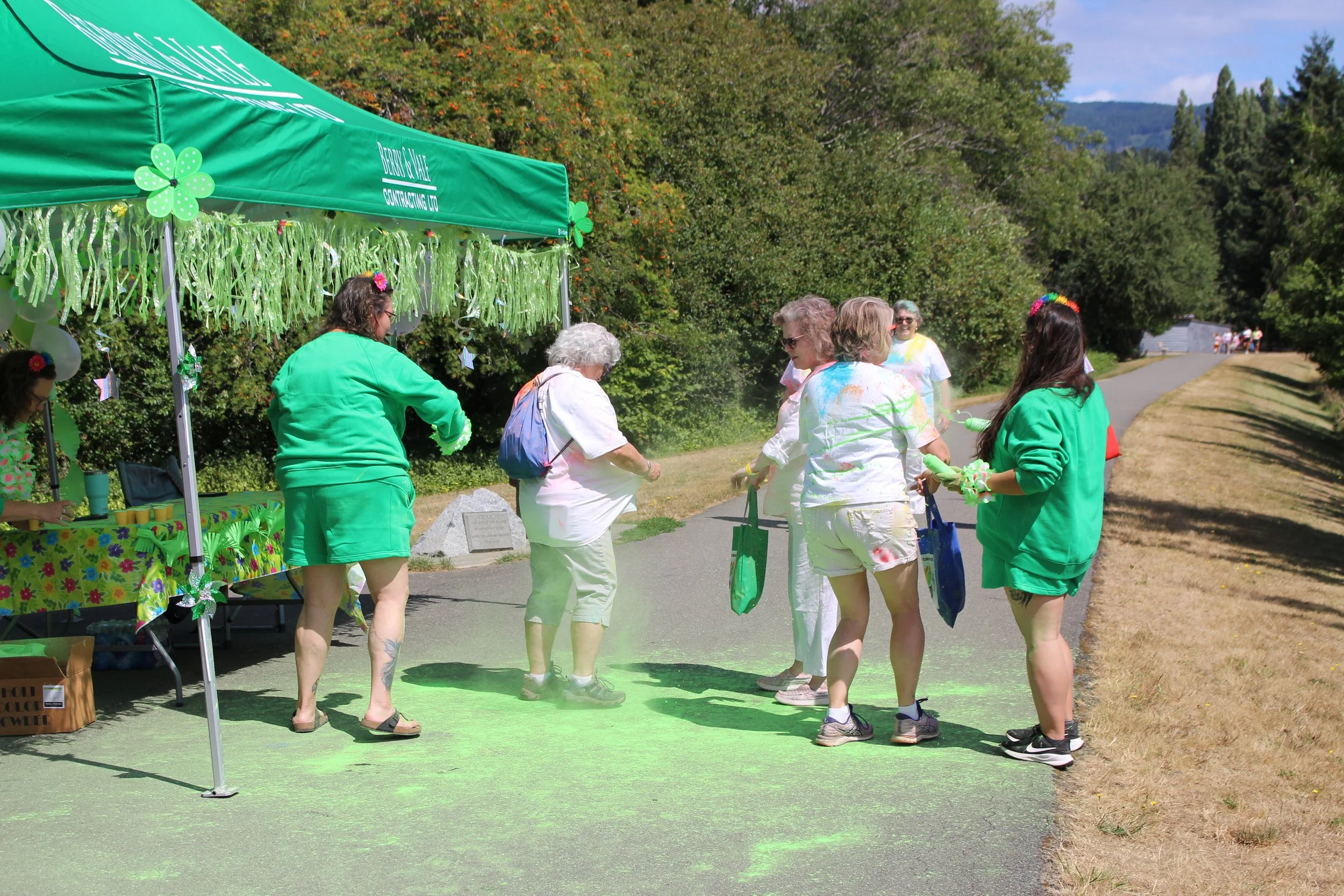 People celebrating during a St. Patrick's Day event, throwing green powder on a paved path outdoors next to a green tent with shamrock decorations, surrounded by trees.