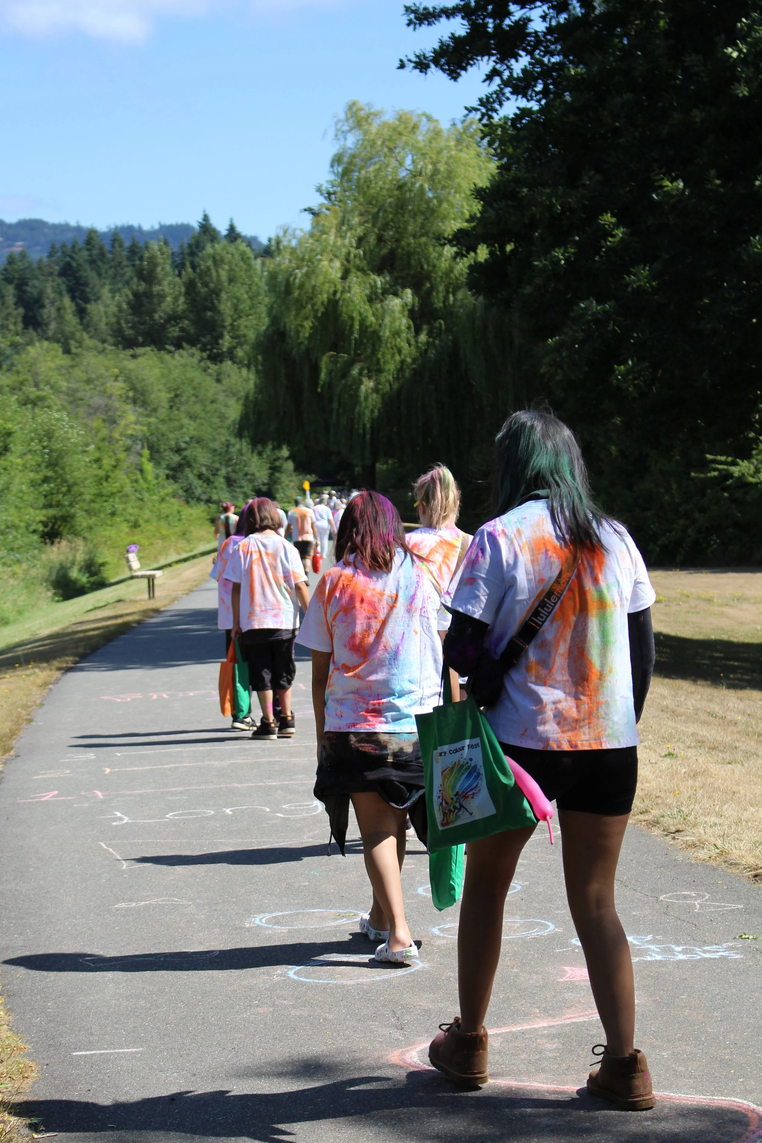 People, mostly young women, walking in a line outdoors on a sunny day, wearing white T-shirts with colorful paint stains, with chalk drawings on the pavement.