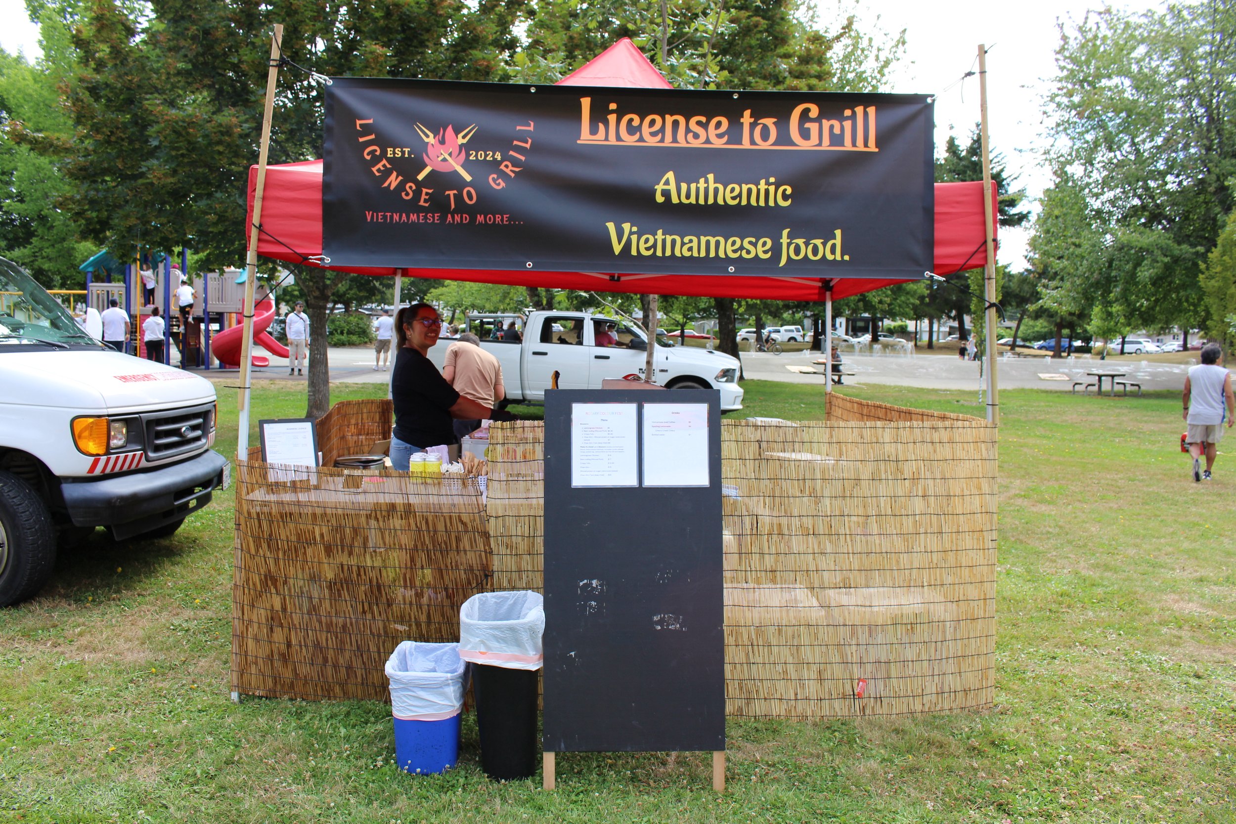 A food stand called 'License to Grill' offering authentic Vietnamese food in an outdoor park. The stand has a black banner with orange and yellow text, bamboo fencing, and a woman behind the counter. There are trash bins in front, and park visitors i