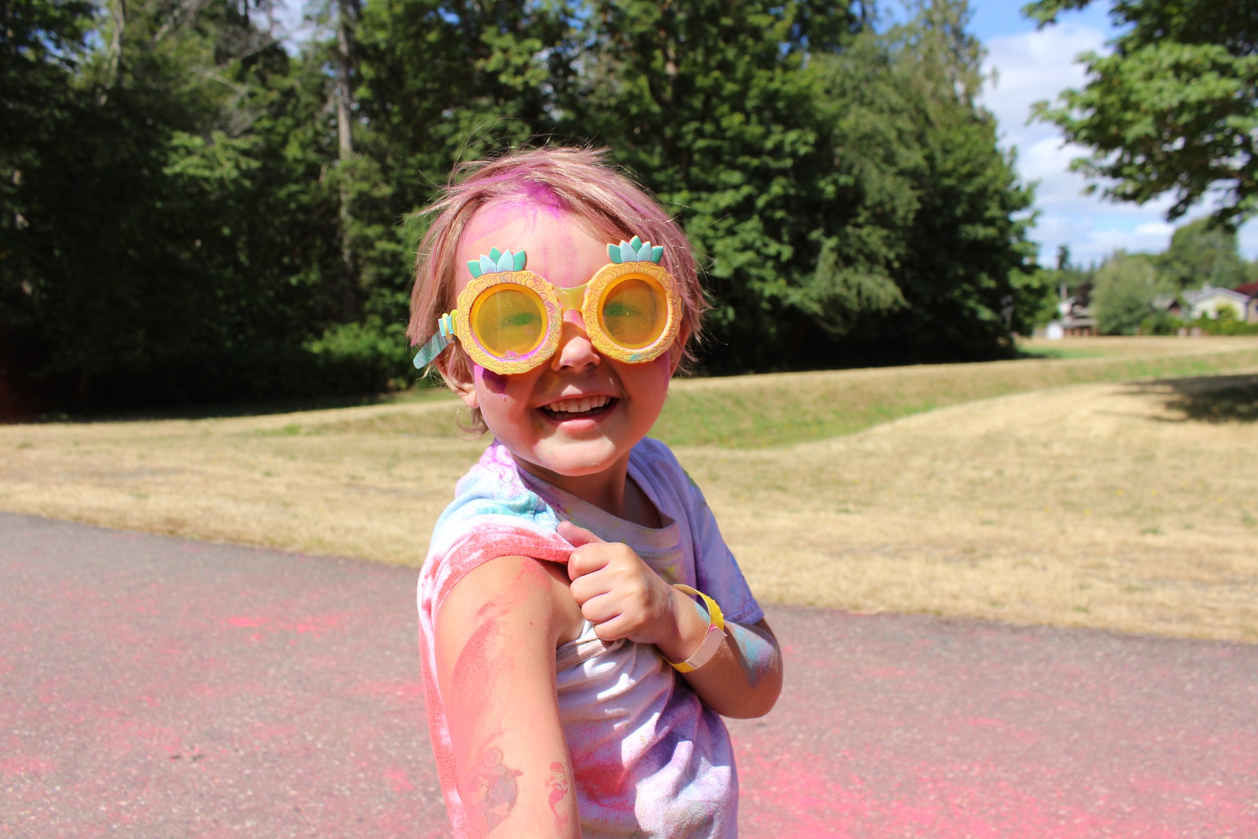 A young girl outdoors wearing large yellow sunglasses with a pineapple design, smiling, with colorful paint on her face and arms, and wearing a tie-dye shirt.
