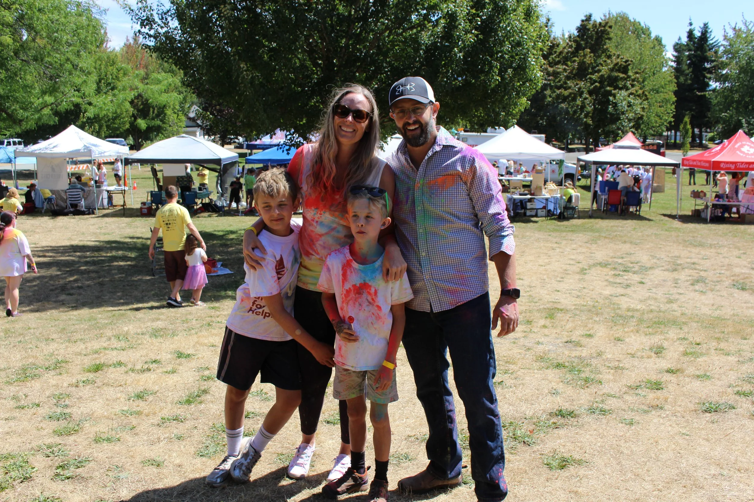 Family of five with two adults and three children standing outdoors at a festival or fair, with tents and other people in the background.