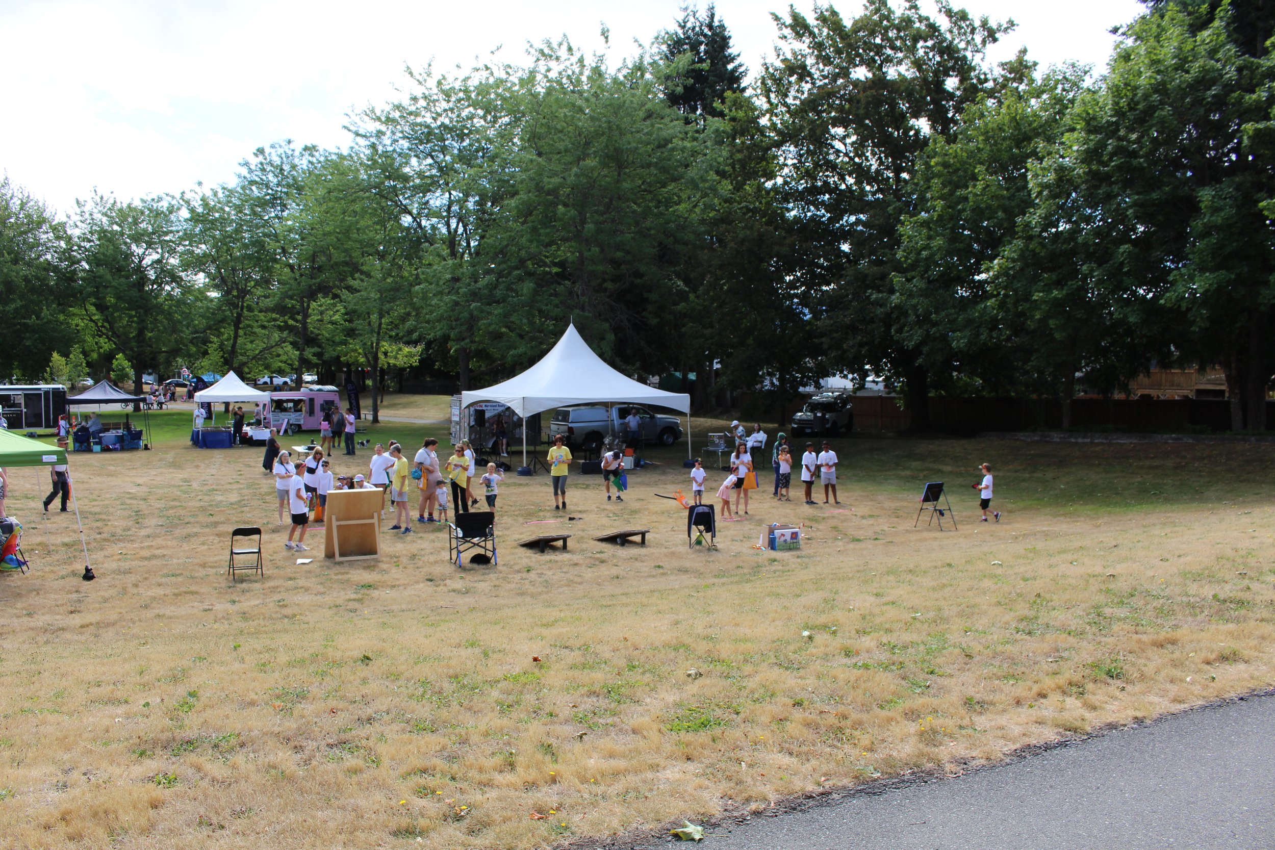 A community outdoor event with tents, chairs, and people gathered in a grassy park area surrounded by trees.