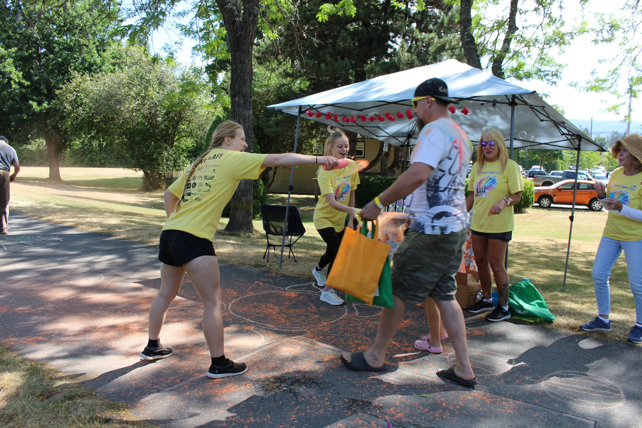 A group of people at an outdoor event, with some wearing yellow shirts, participating in a game or activity involving a small flame or torch, under a tent on a sunny day.