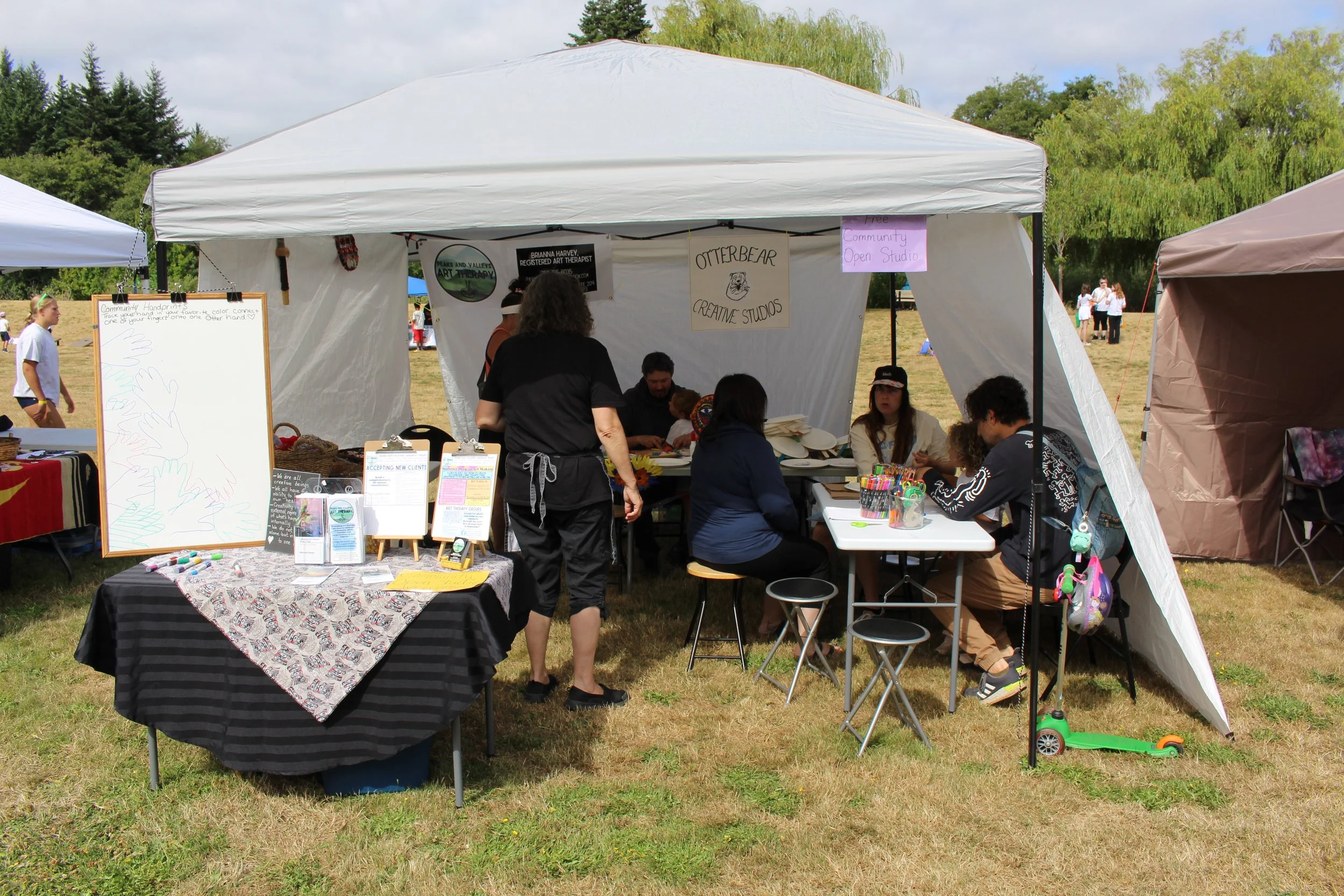 Community art booth with people creating and displaying art at an outdoor fair, with signs indicating free community open studio, and art supplies on tables.