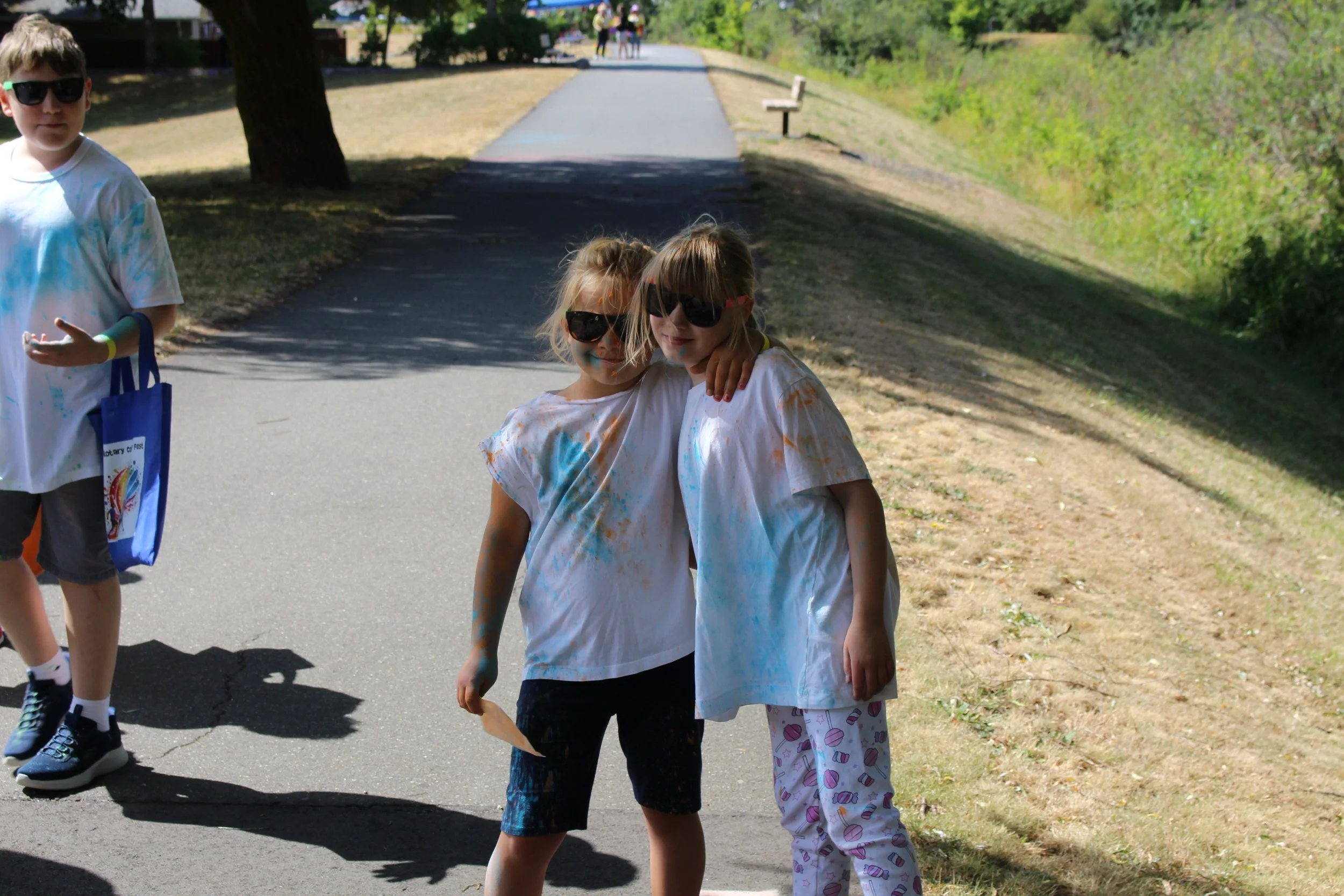 Two young girls wearing oversized sunglasses and white t-shirts with colorful paint splatters, standing together outside on a sunny day, hugging and smiling, with a boy nearby, also dressed in white with paint, in a park or outdoor recreational area.