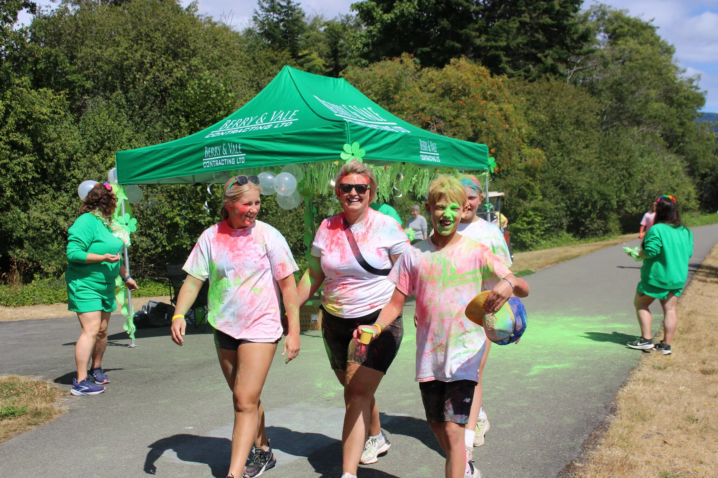 People participating in a color run, covered in colorful powder, walking on a park trail near a green tent with balloons and decorations.
