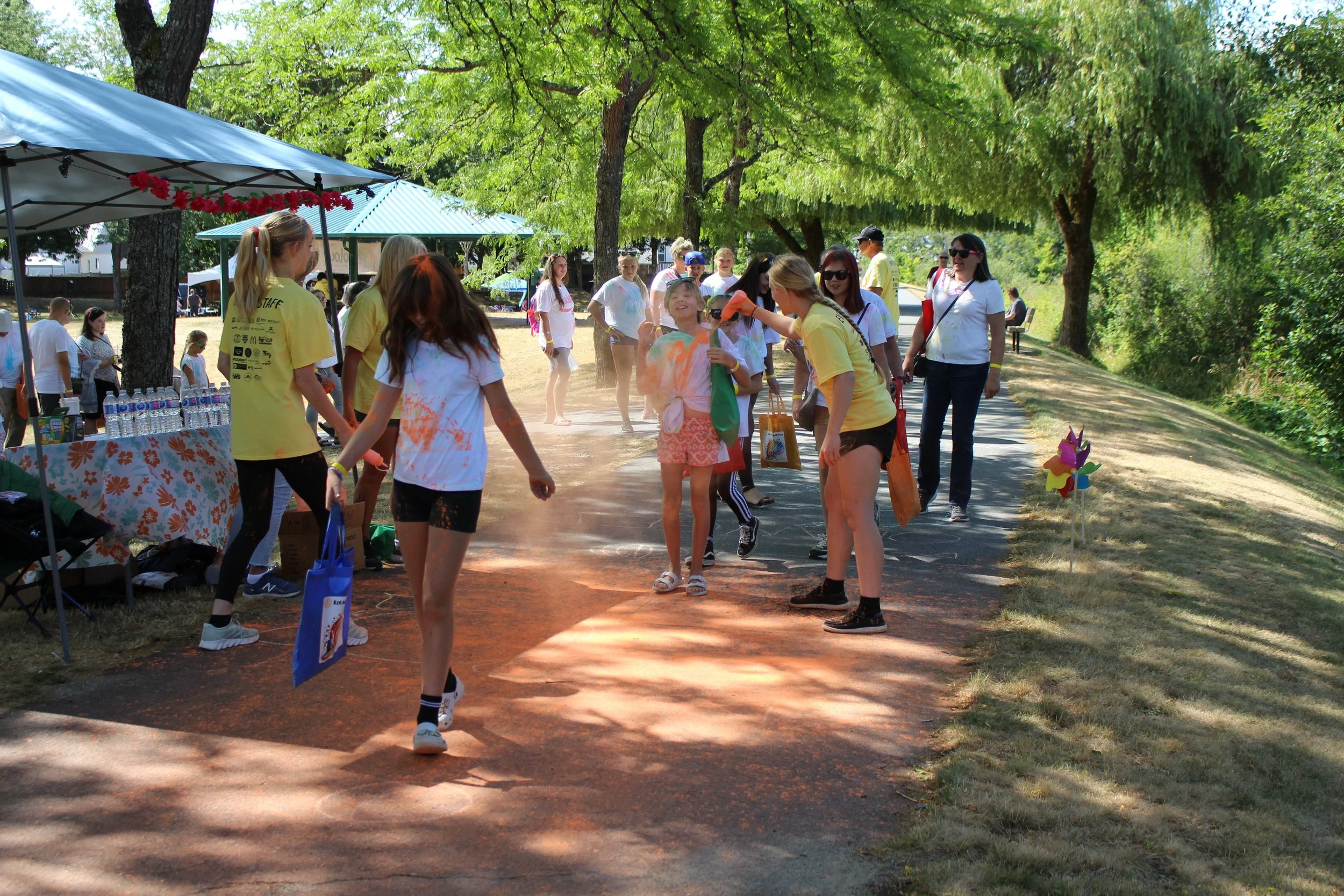 People participating in a color run, throwing colored powder at each other outdoors on a sunny day with trees and tents in the background.