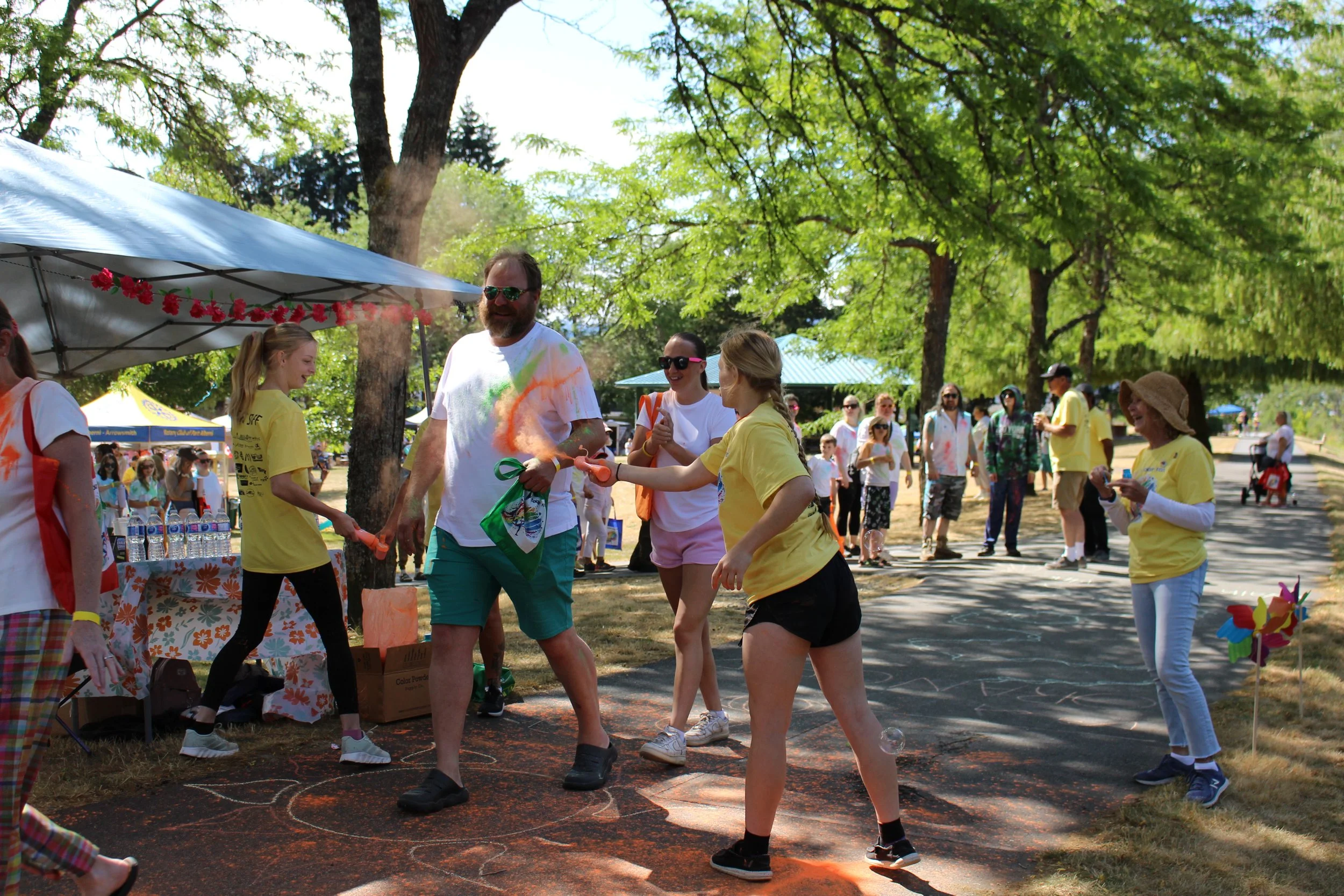 People at an outdoor festival or event, some wearing yellow shirts, being playful and spraying colorful powder on each other under green trees with a blue sky.
