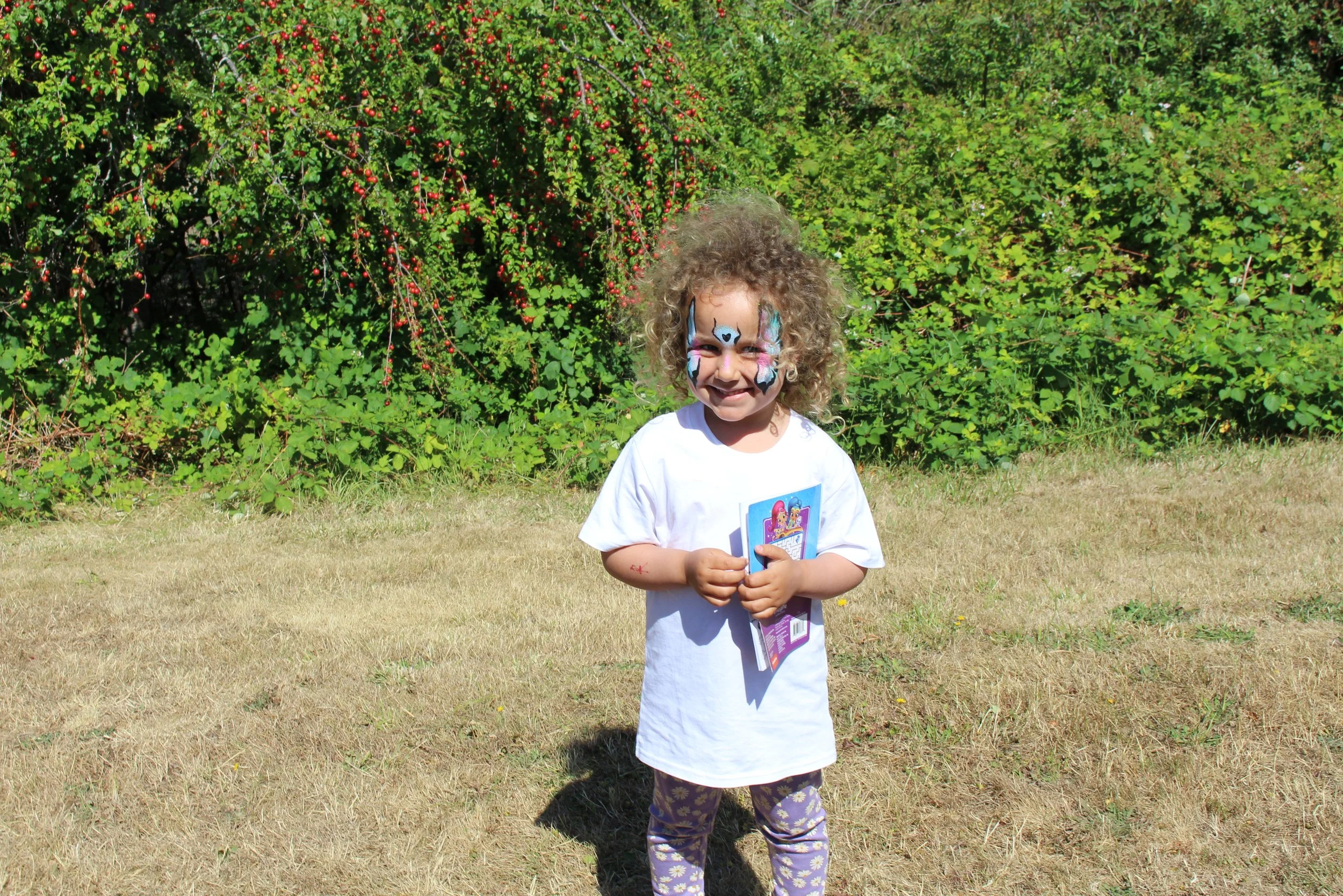 A young girl with face paint of a dog and pink and blue accents, wearing a white t-shirt and purple patterned leggings, standing outdoors on a grassy area with tall green bushes in the background, smiling and holding a booklet.