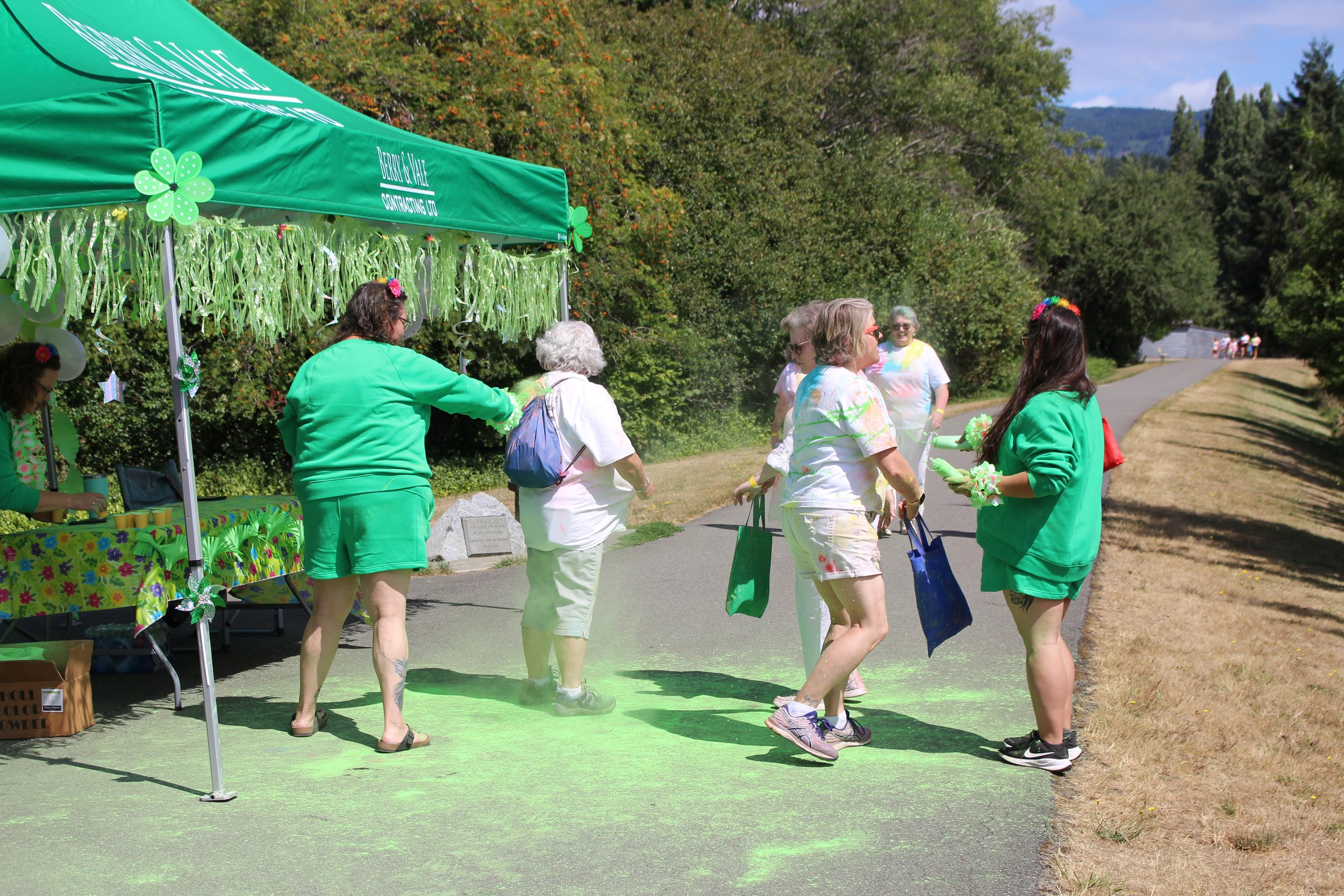 People participating in a color run event outdoors, throwing green powder, near a green tent with decorations.