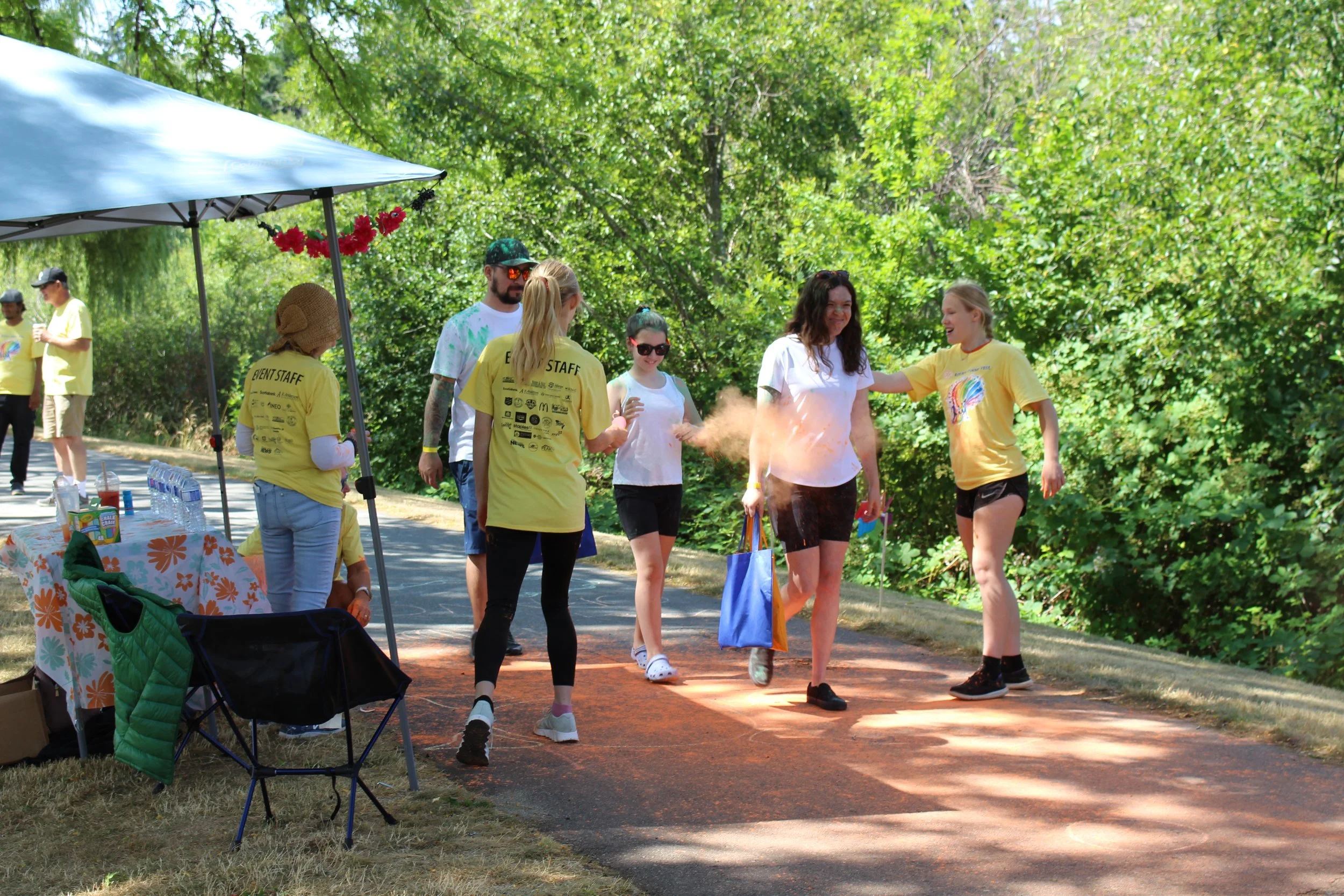People participating in a color powder run event, with one person covered in orange powder, under a canopy on a path surrounded by green trees.