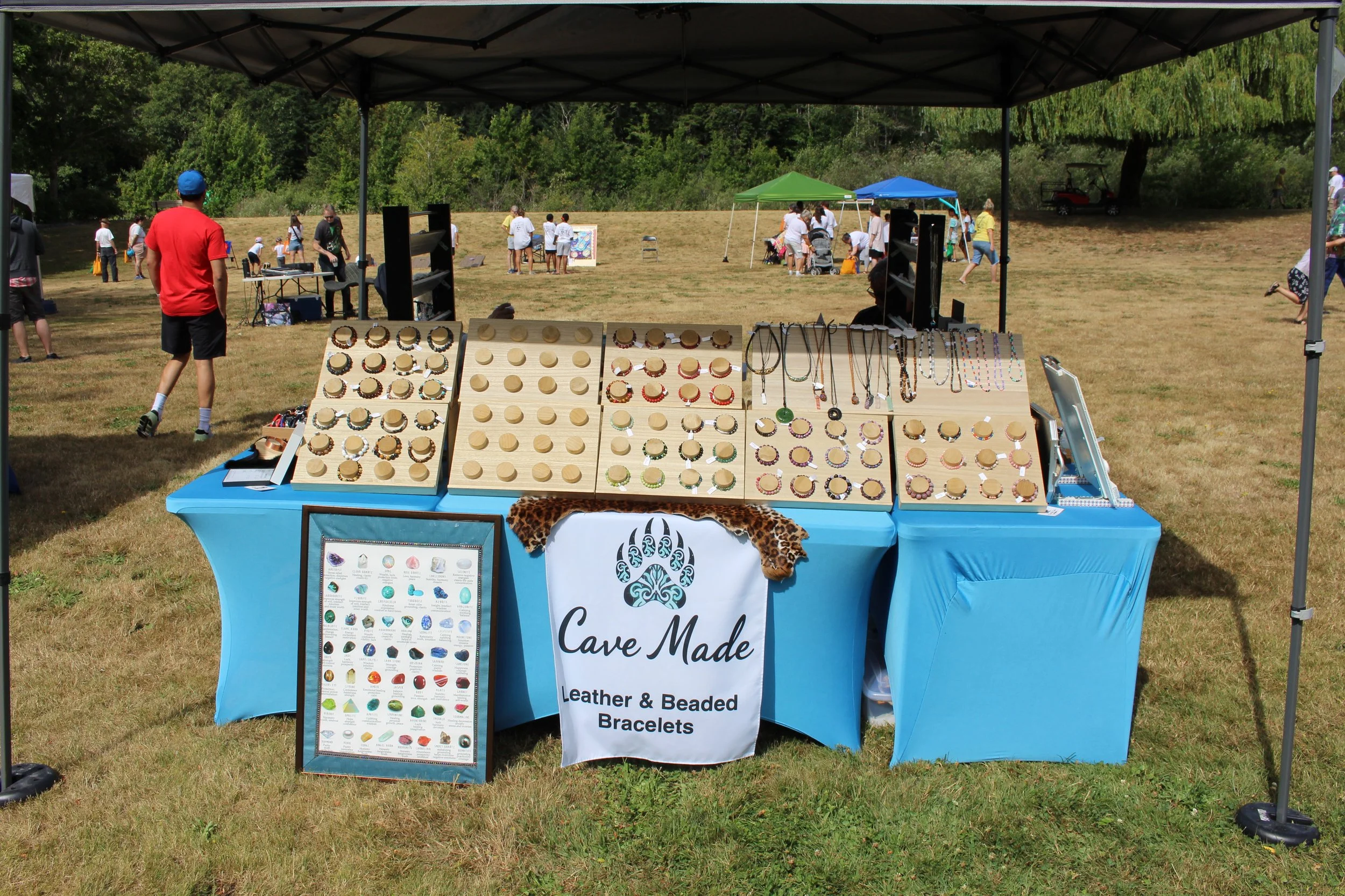 Outdoor jewelry stand with leather and beaded bracelets on display at a craft fair, with people and tents in a park setting.