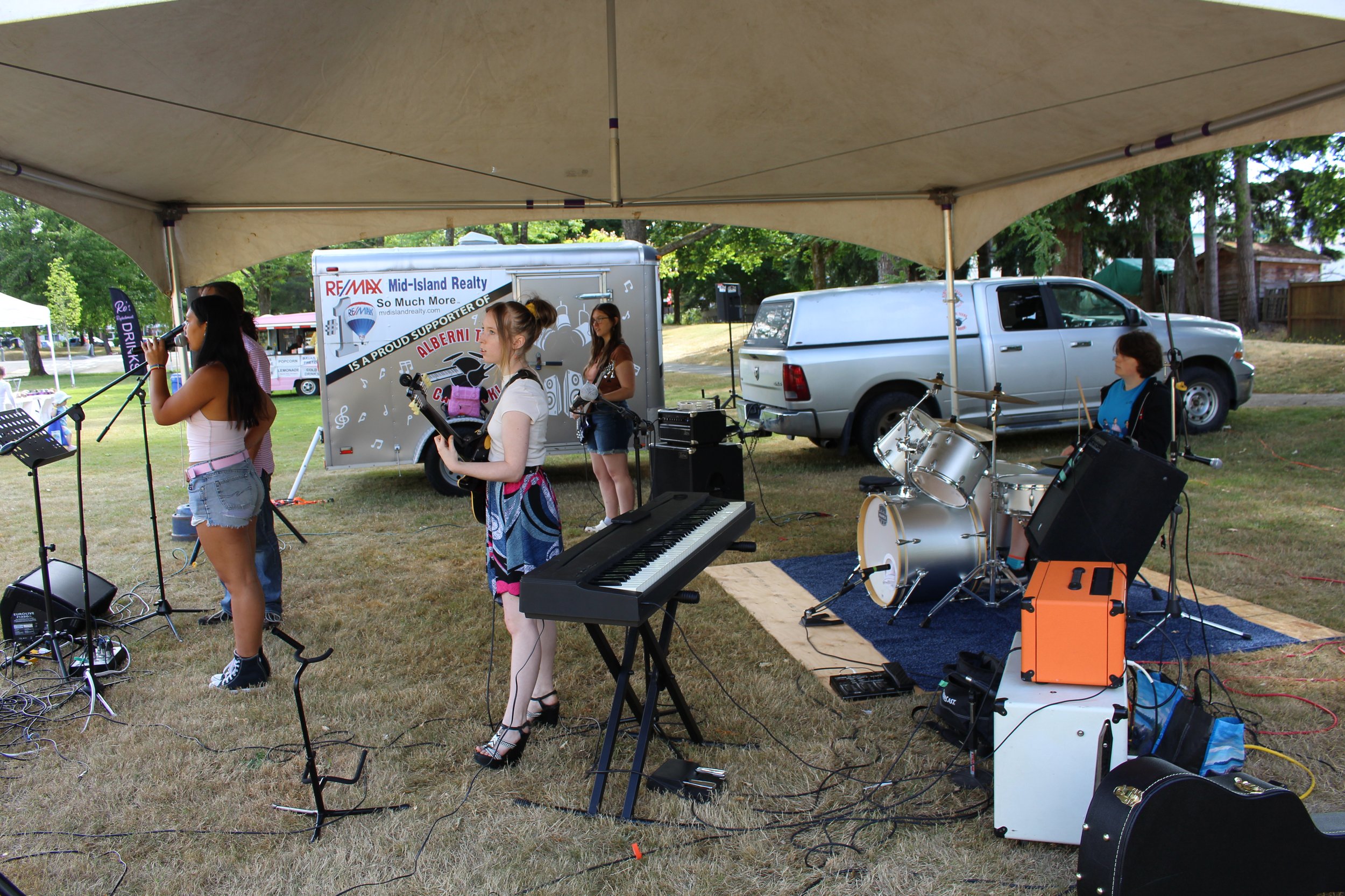 A band performs on stage at an outdoor event, with young women playing instruments such as guitar and drums under a tent, and trucks and trees in the background.