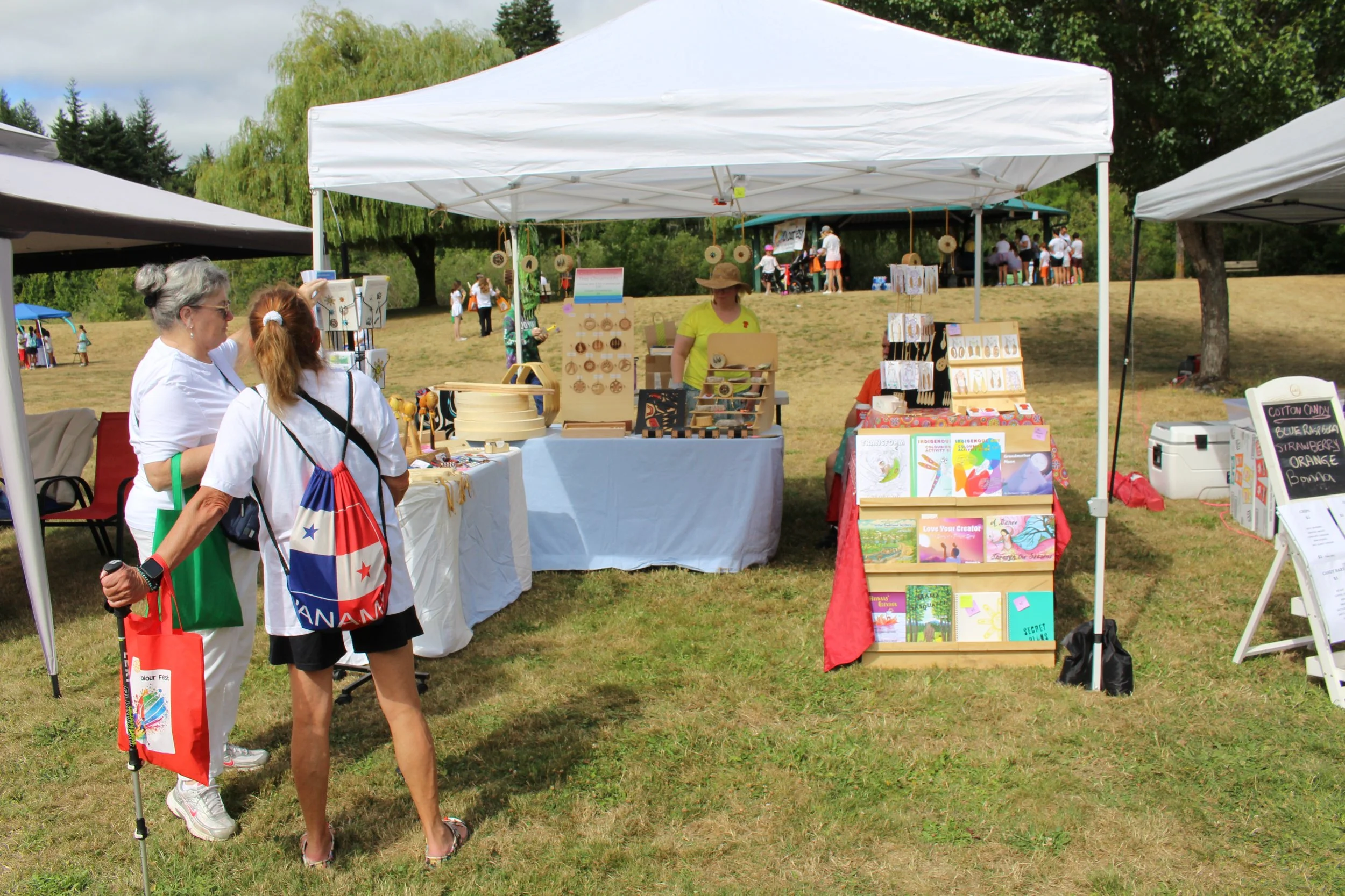 People shopping at outdoor craft market stalls on grassy field under tents with trees in the background.