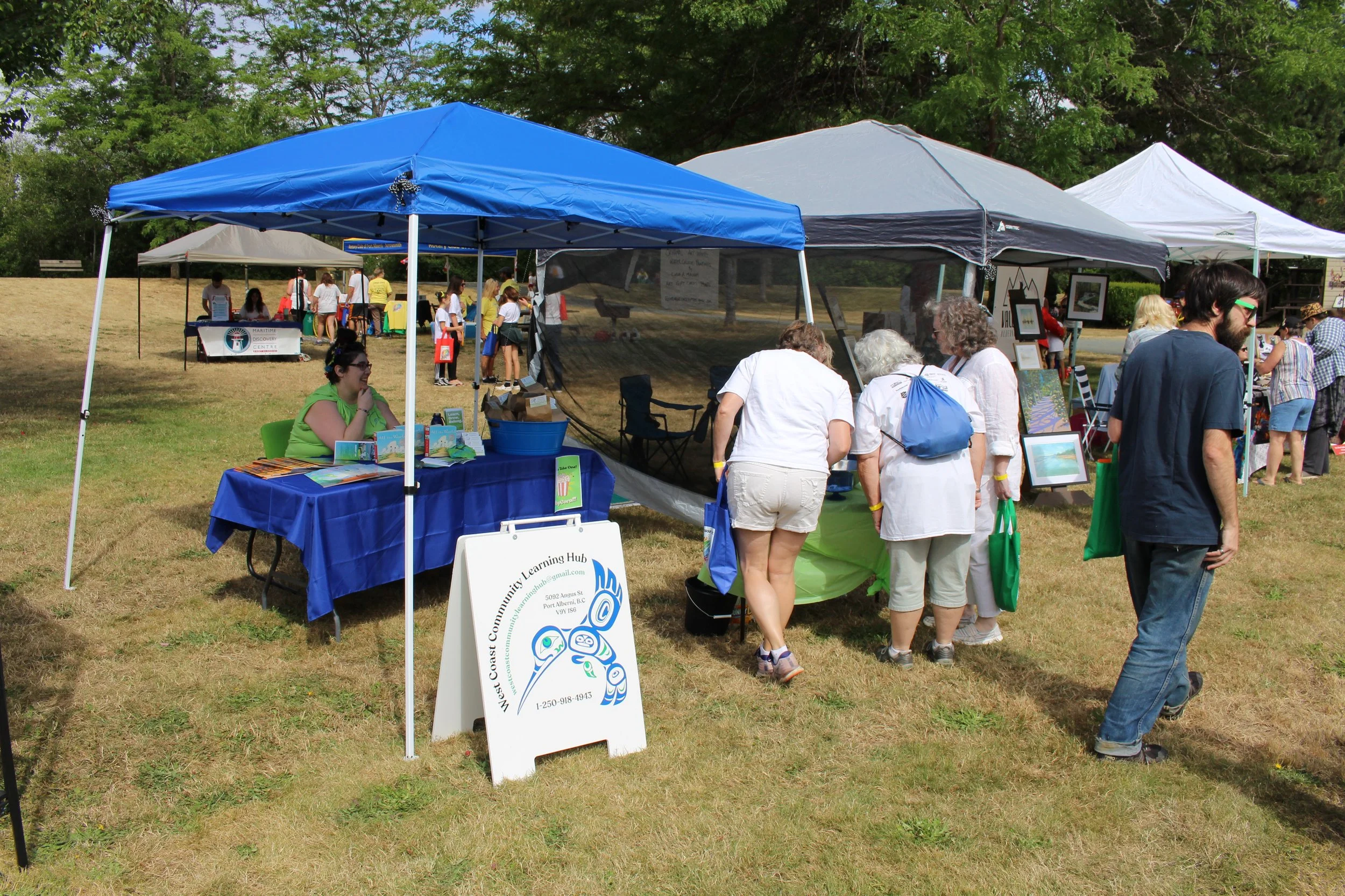Outdoor community event with several tents, people browsing booths, and a sign for Western Coast Community Learning Hub in a grassy area with trees.