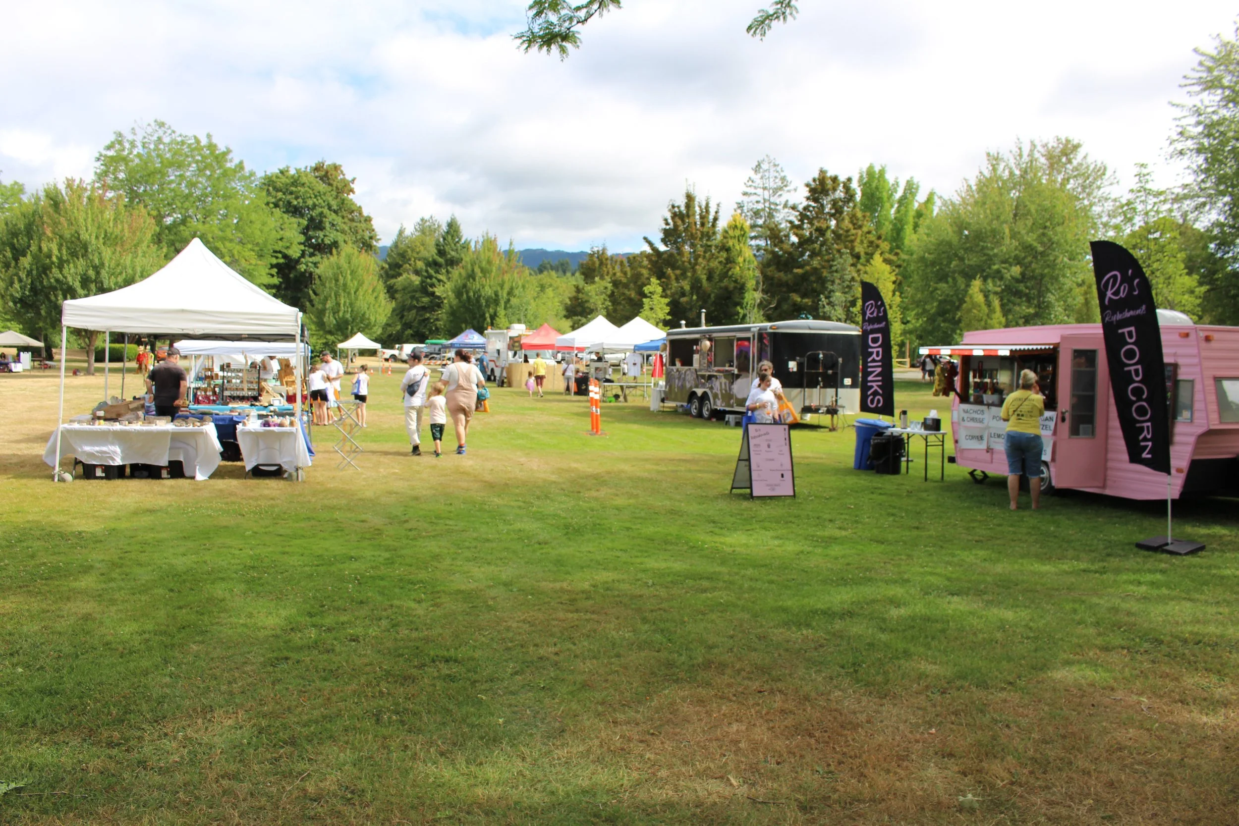 Outdoor fair with food trucks, vendor booths, and visitors on a grassy field with trees in the background.