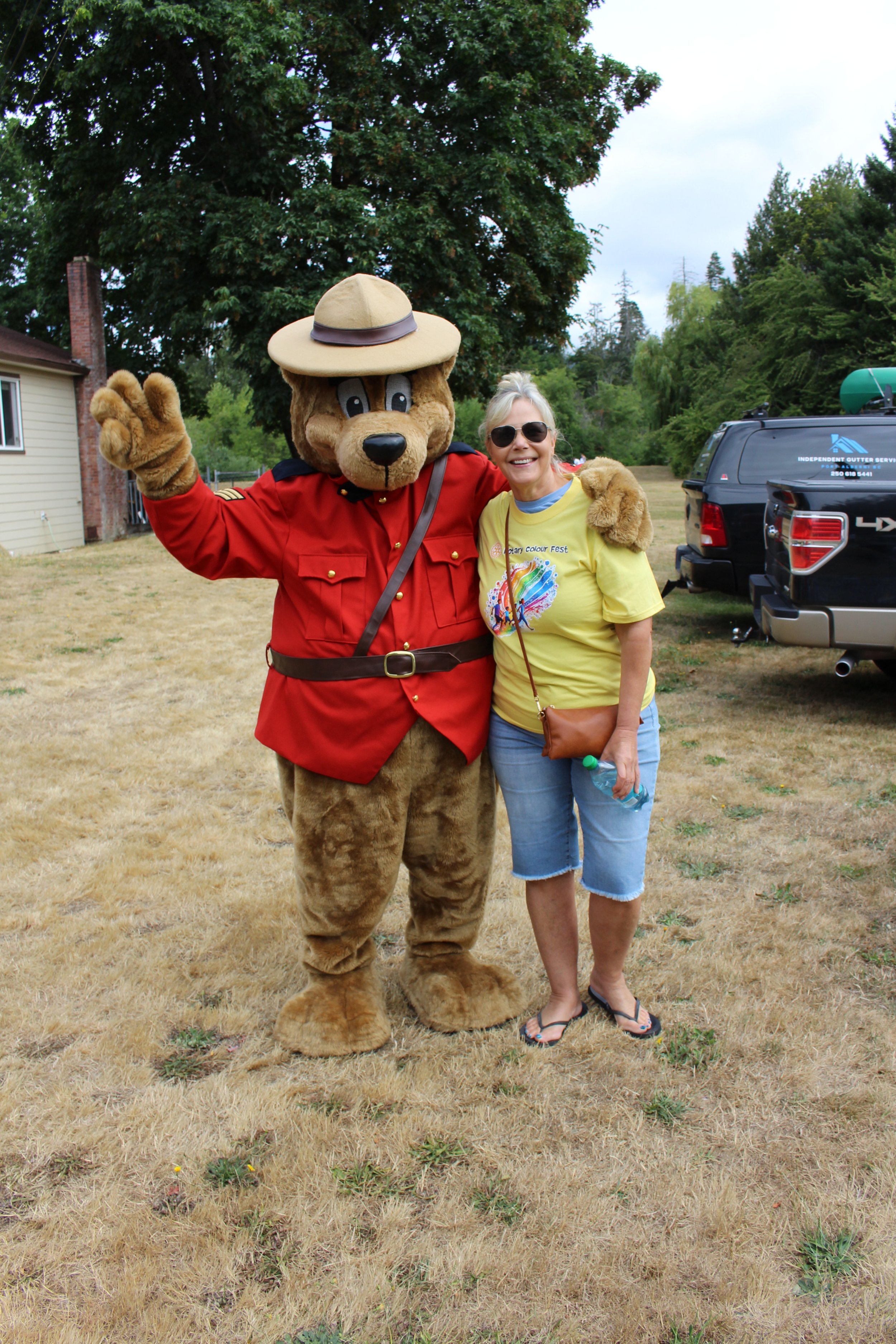 A woman wearing sunglasses and a yellow T-shirt stands next to a person in a bear mascot costume dressed as a park ranger, both smiling and posing outdoors on a grassy area with trees, houses, and parked cars in the background.