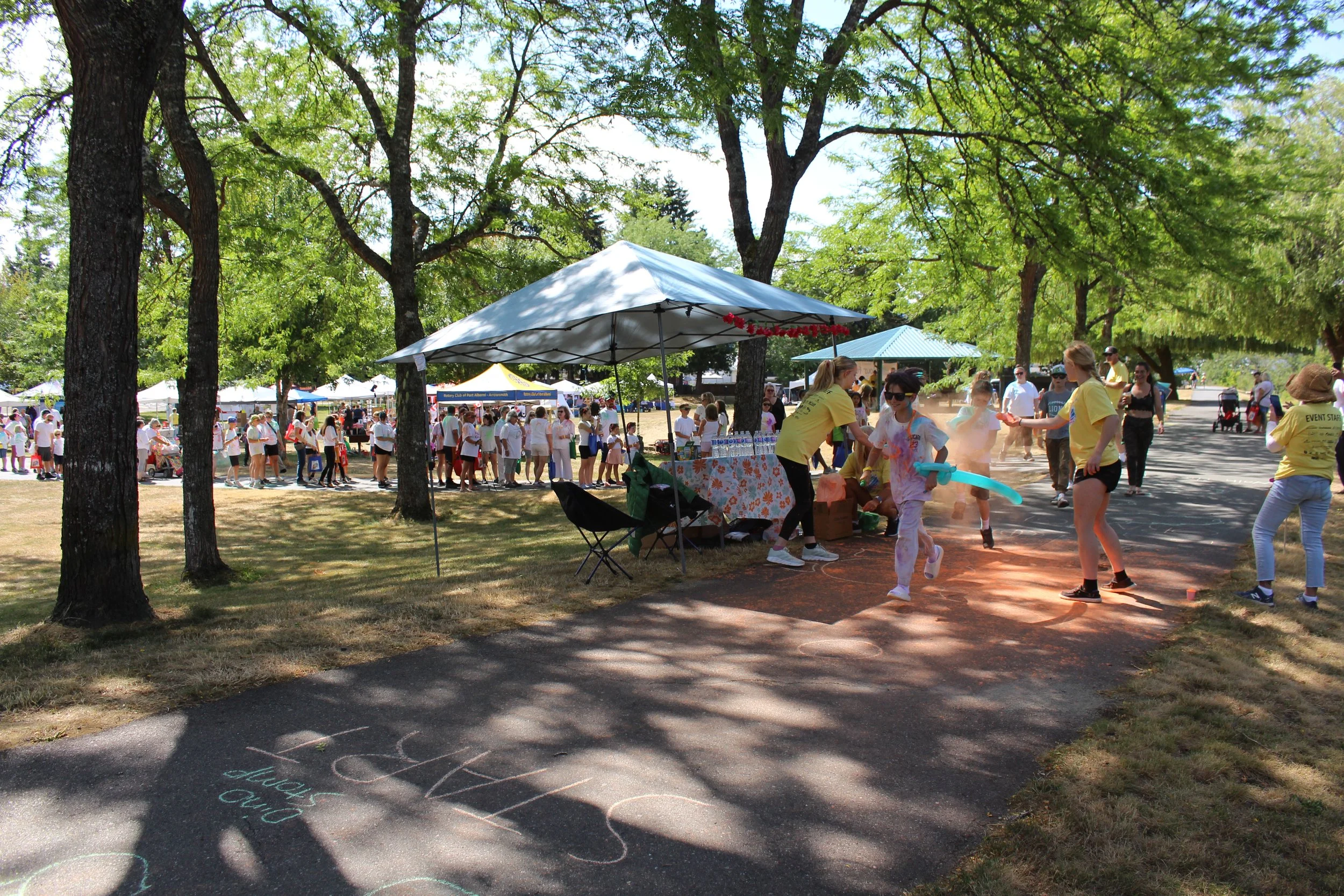 Children running and playing with water balloons at an outdoor event in a park with many people, tents, and shaded trees.