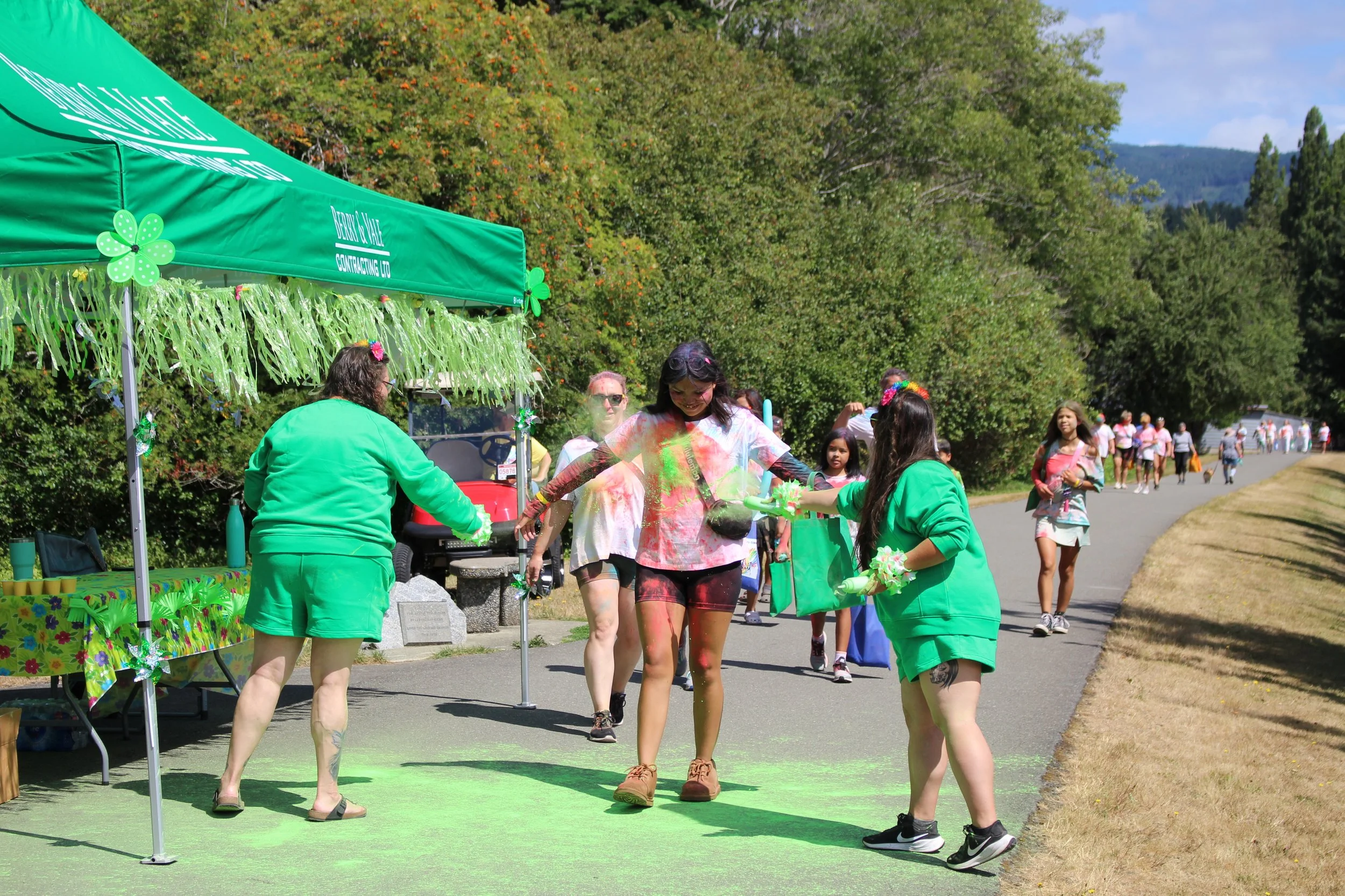 People celebrating Holi with colorful powders in a park, some on a green grass mat, others walking by.