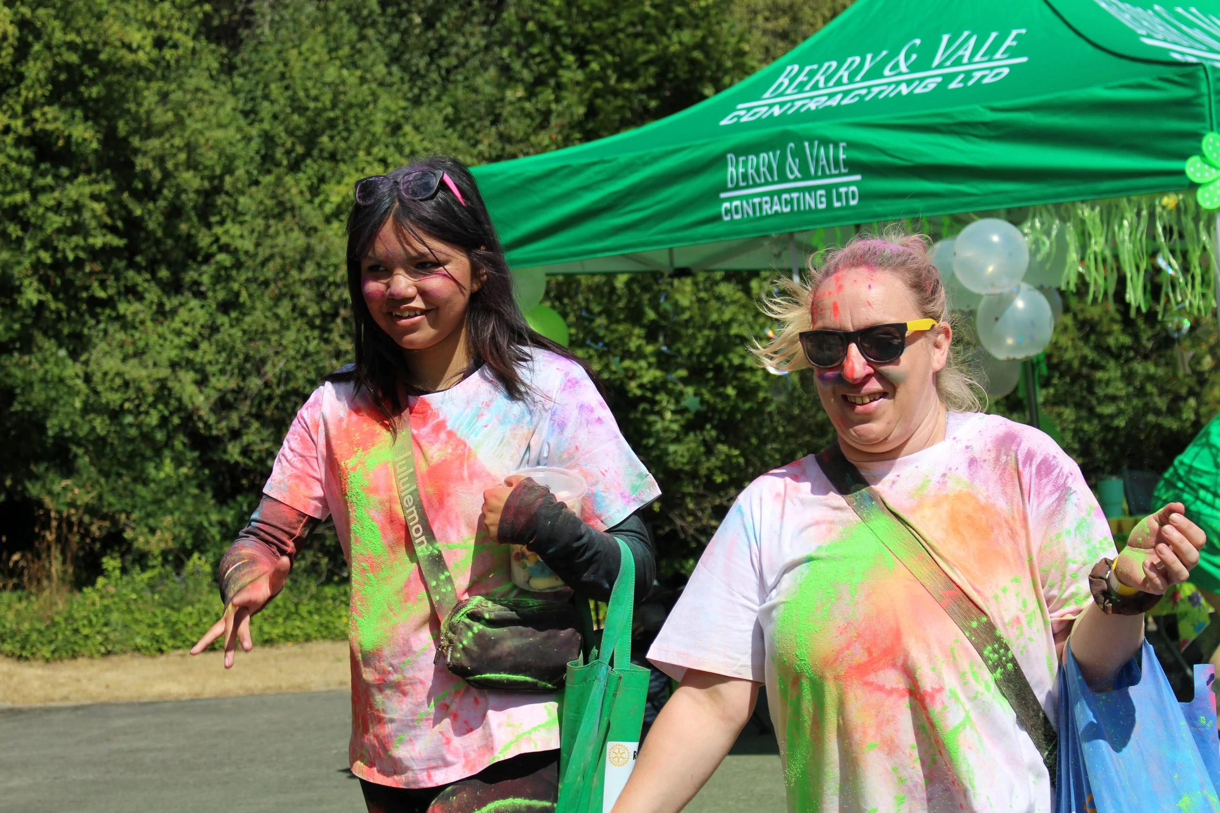 Two smiling women covered in colorful powder at an outdoor event, with a green pennant tent reading 'Berry & Vale Contracting LTD' behind them, and balloons decorating the tent.