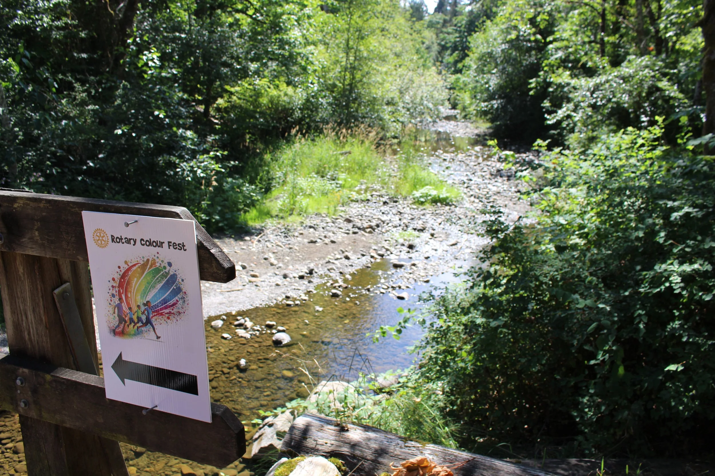 A wooden sign for the Rotary Colour Fest with a rainbow design and silhouettes of people, located on a bridge overlooking a shallow stream surrounded by green trees and bushes on a sunny day.