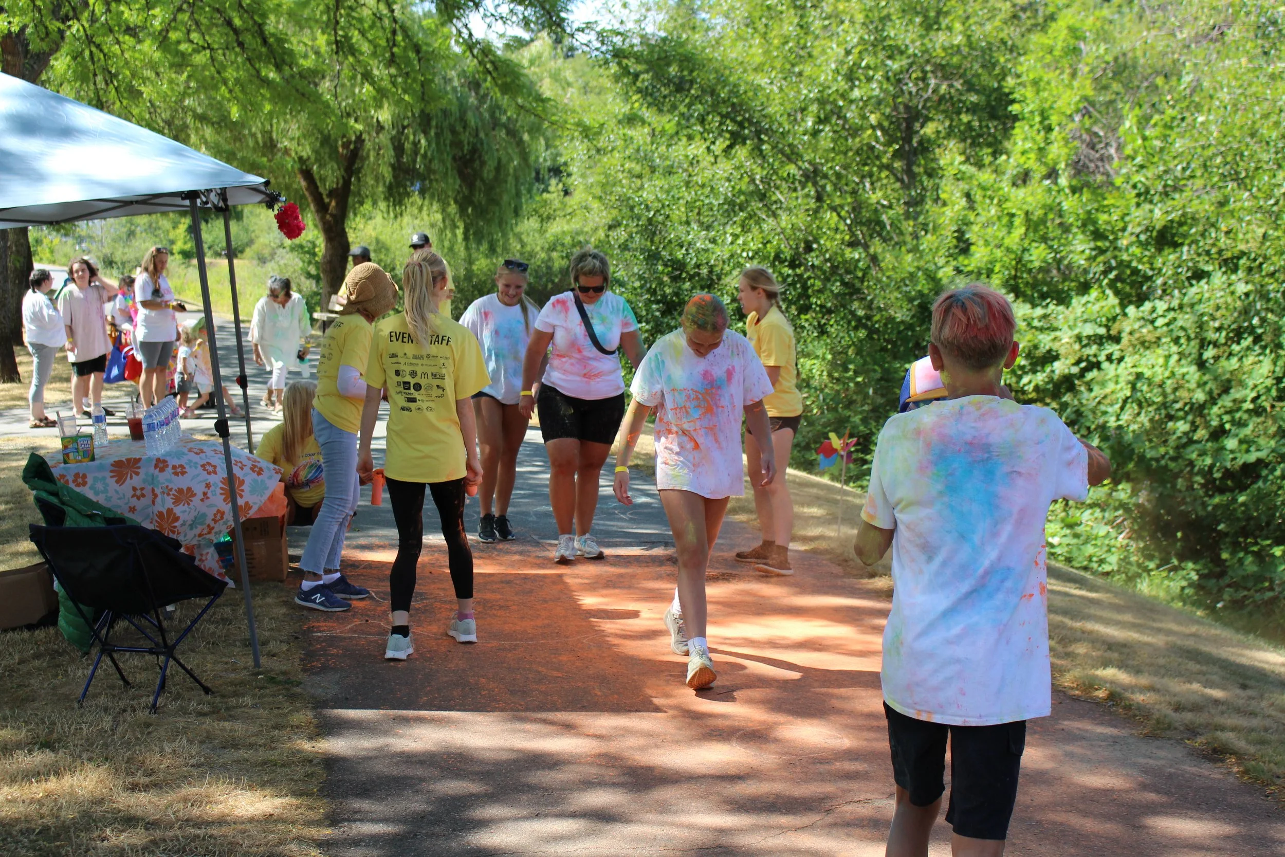People participating in a color run event outdoors, wearing white shirts with colorful powder on them, in a park with green trees.