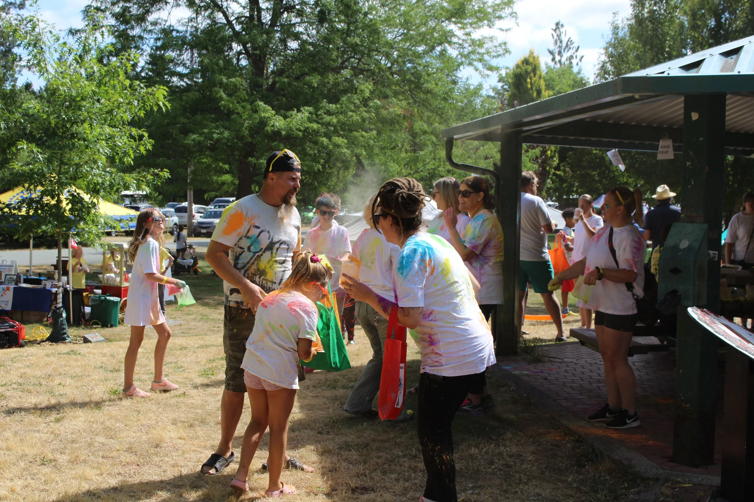People celebrating a color festival outdoors, covered with colorful powder, engaging in an activity near a pavilion, with trees and parked cars in the background.