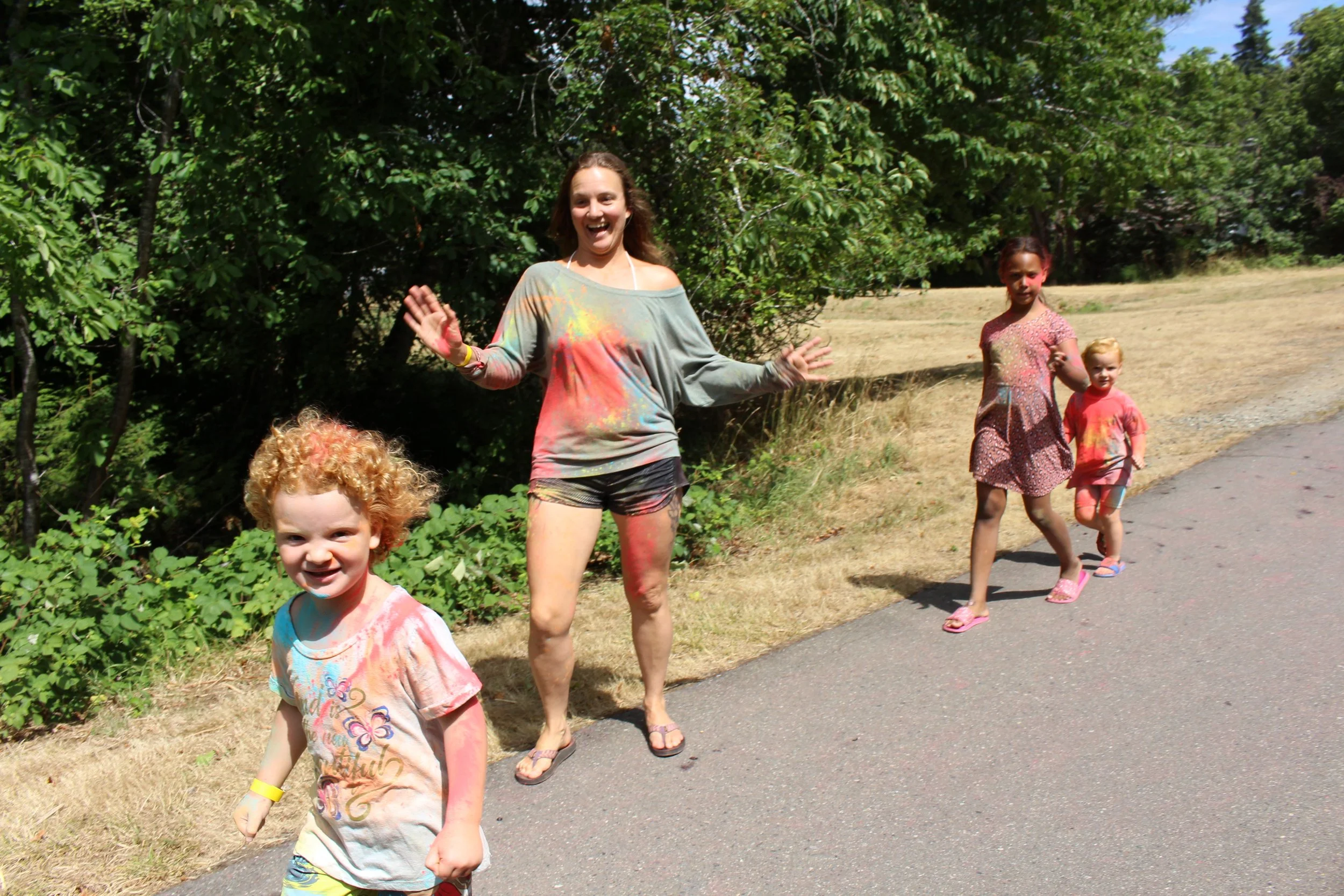 Four children and one woman walking along a paved path outdoors, covered in colorful powder, likely celebrating a festival like Holi, with trees and grass in the background.