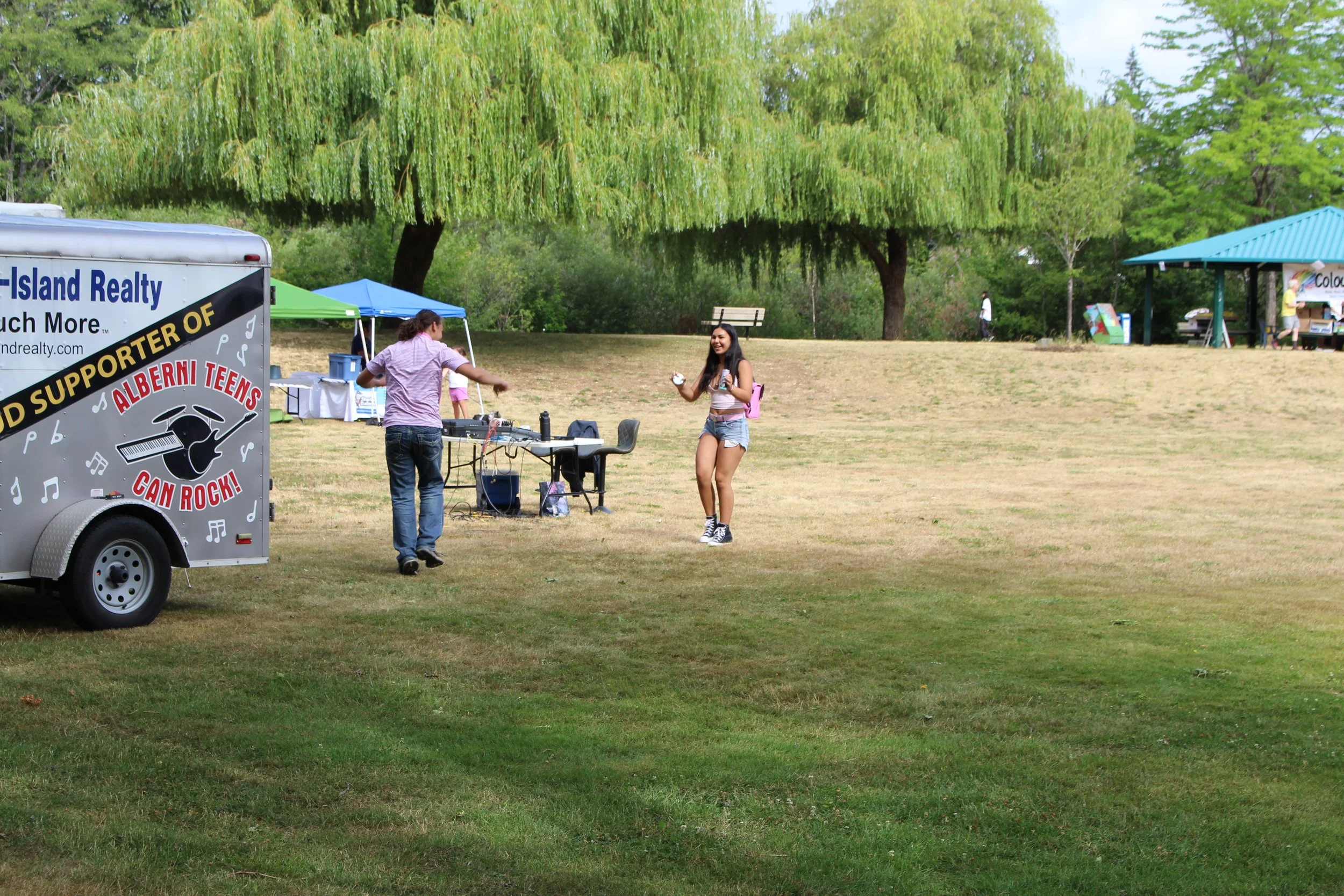 A young woman dancing at an outdoor park event with a man DJing from a mobile setup. There are tents and a support sign for Alberni Teens Can Rock. Trees and a pavilion are in the background.