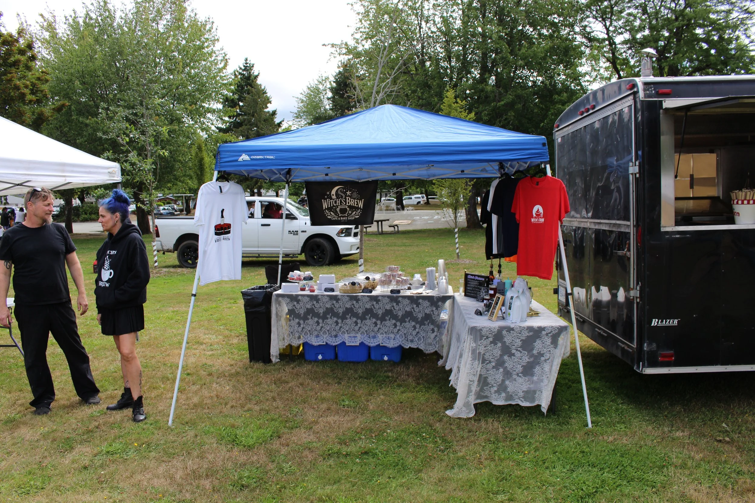 Outdoor market stall with Halloween-themed merchandise, ice cream, and baked goods, set up on grass with trees in the background.