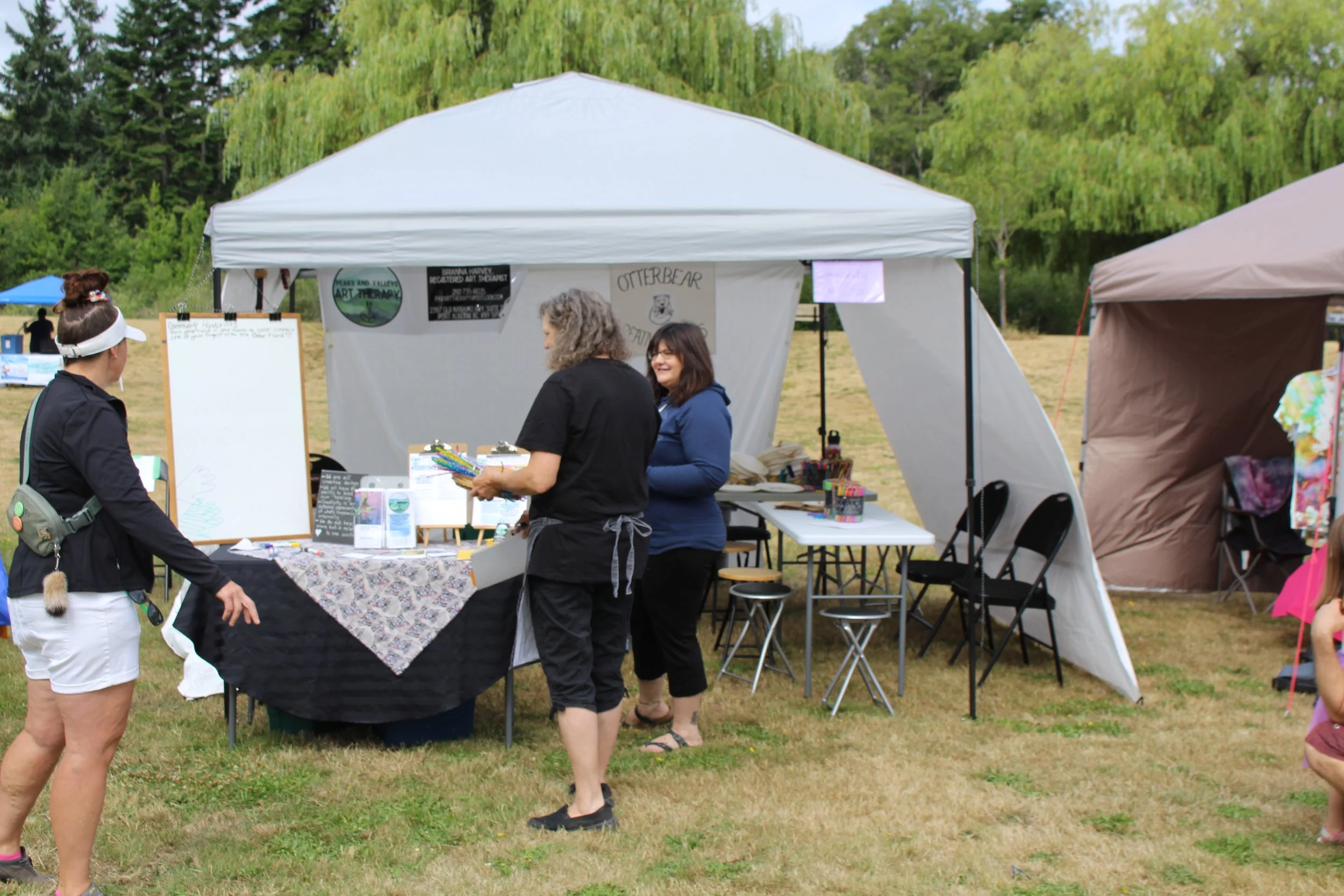 People at an outdoor craft fair booth with art supplies and signs, one person browsing and two vendors behind the table.