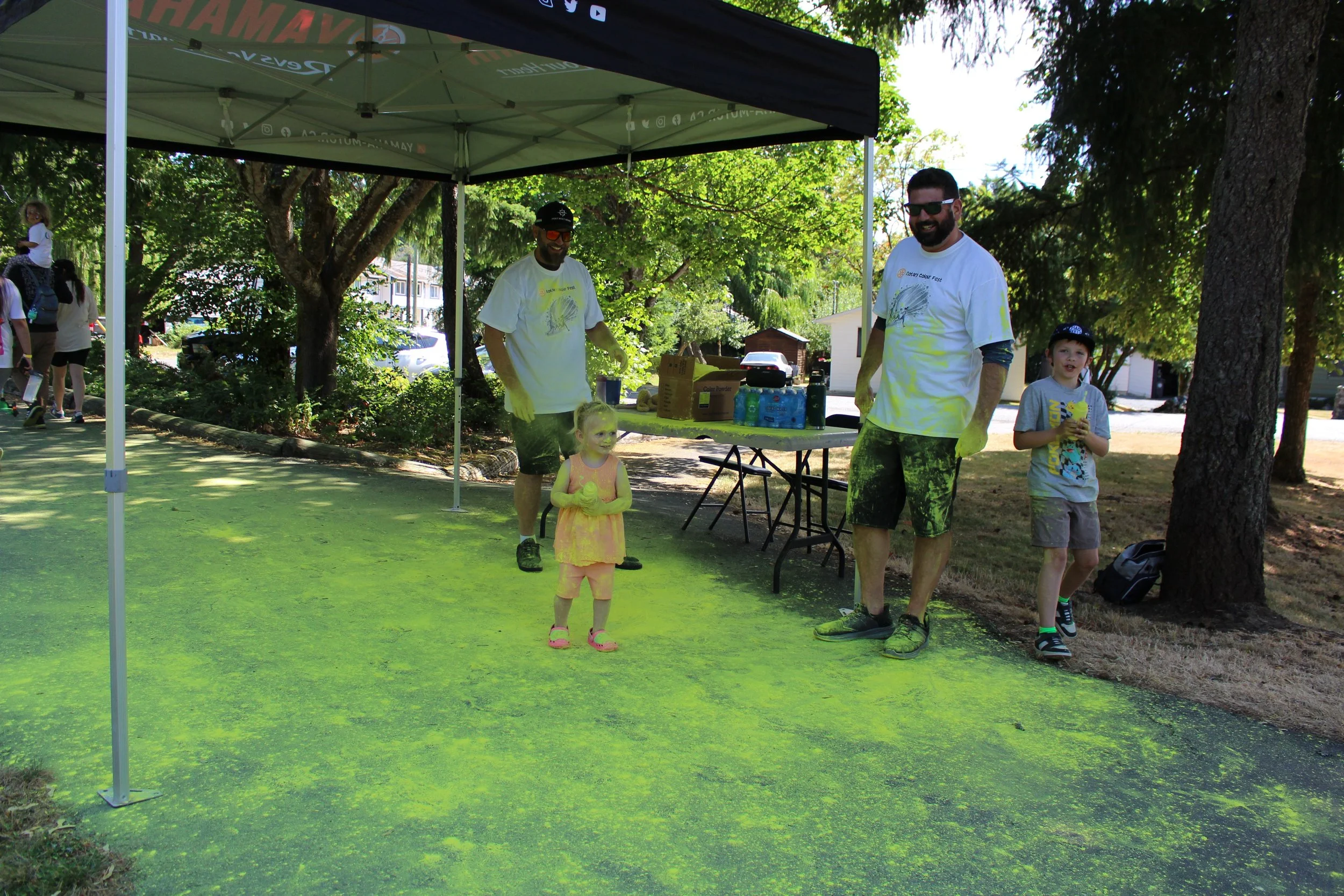 Four people, two adults and two children, under a green canopy at a park, with yellow powder on the ground and on their clothes, participating in a color run event.
