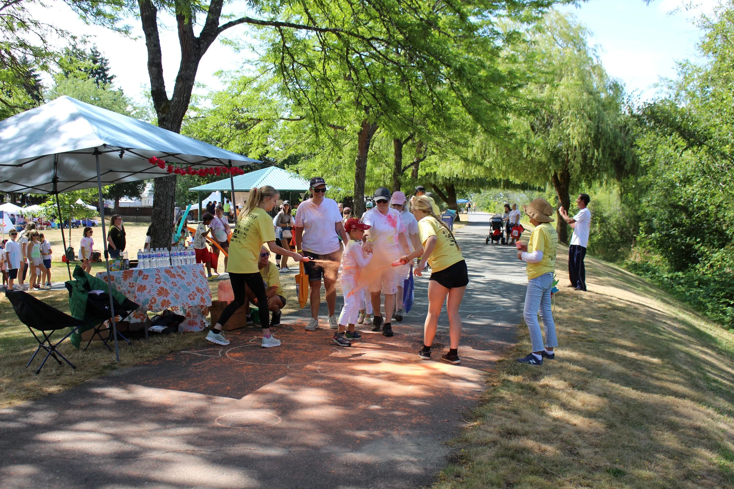 People at an outdoor event playing a game where they toss powder onto each other, surrounded by trees and tents, on a sunny day.