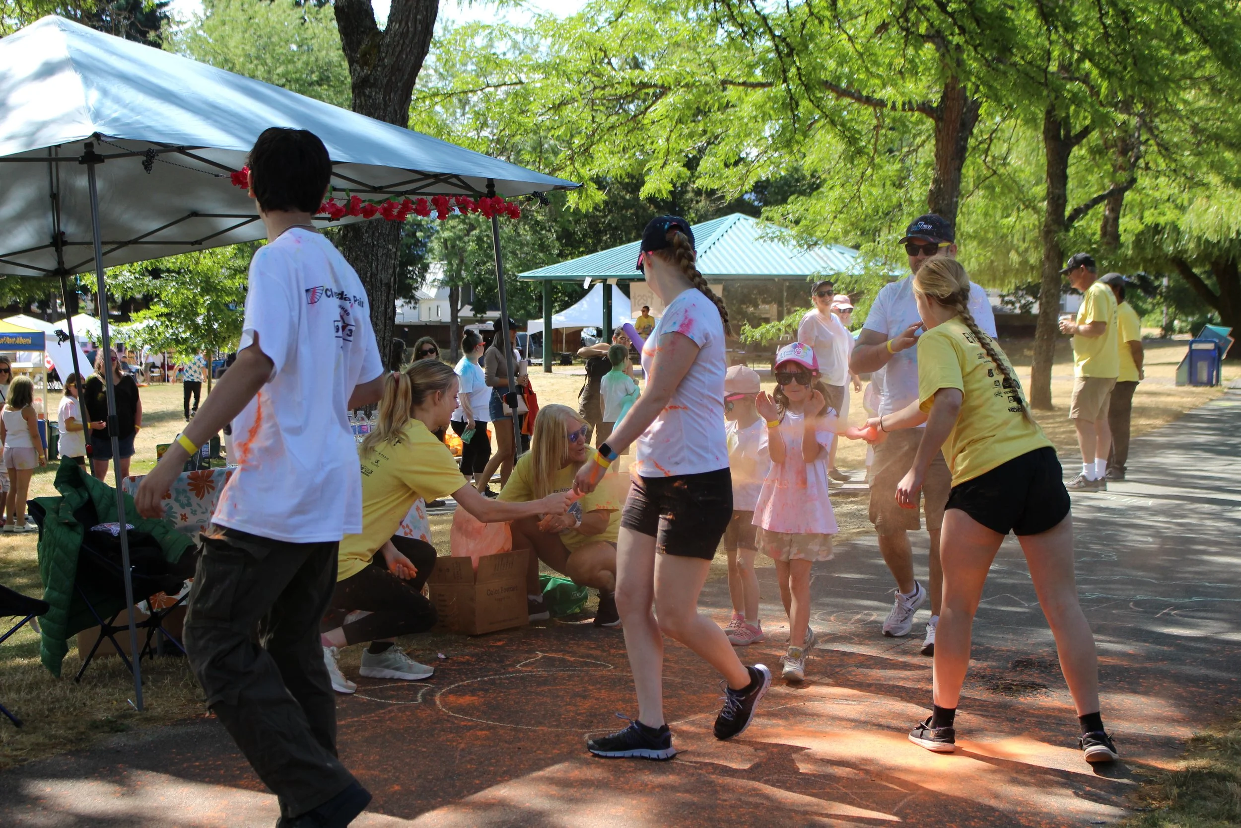 People celebrating at an outdoor event with color powder, children and adults laughing, running, and throwing colored powder in a park with trees and tents.