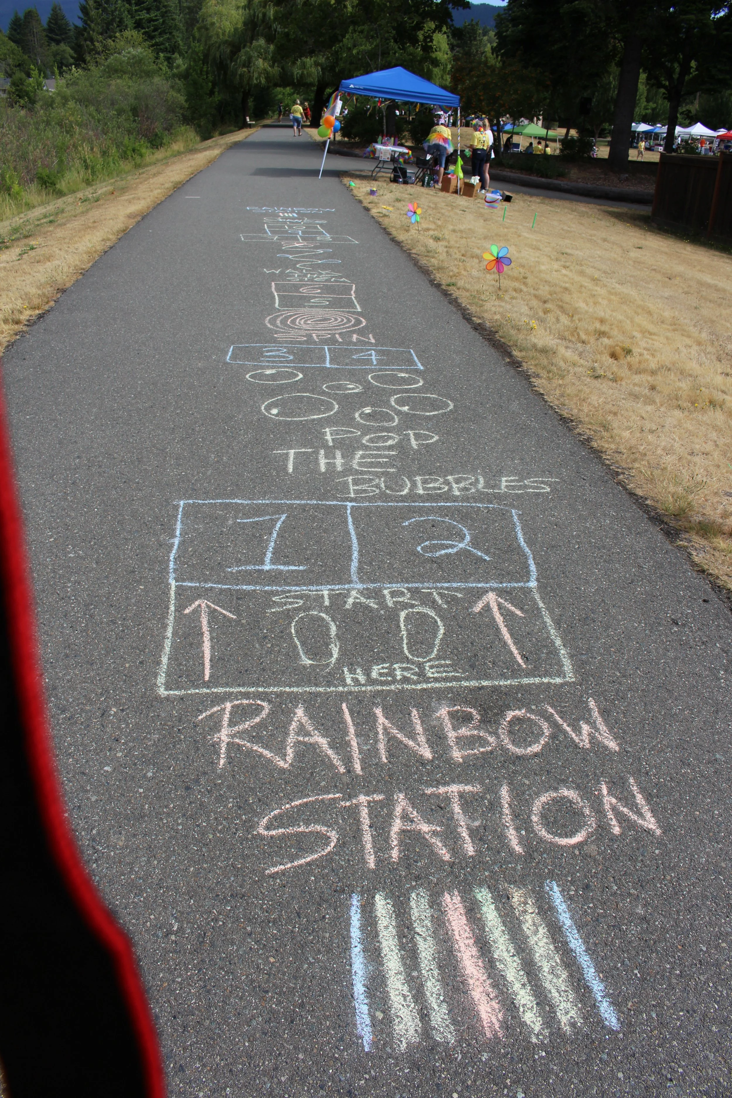 Colorful chalk hopscotch game drawn on pavement with a rainbow station sign at the start and a sign for Rainbow Station on the pavement. Tents and people in the background at an outdoor event.