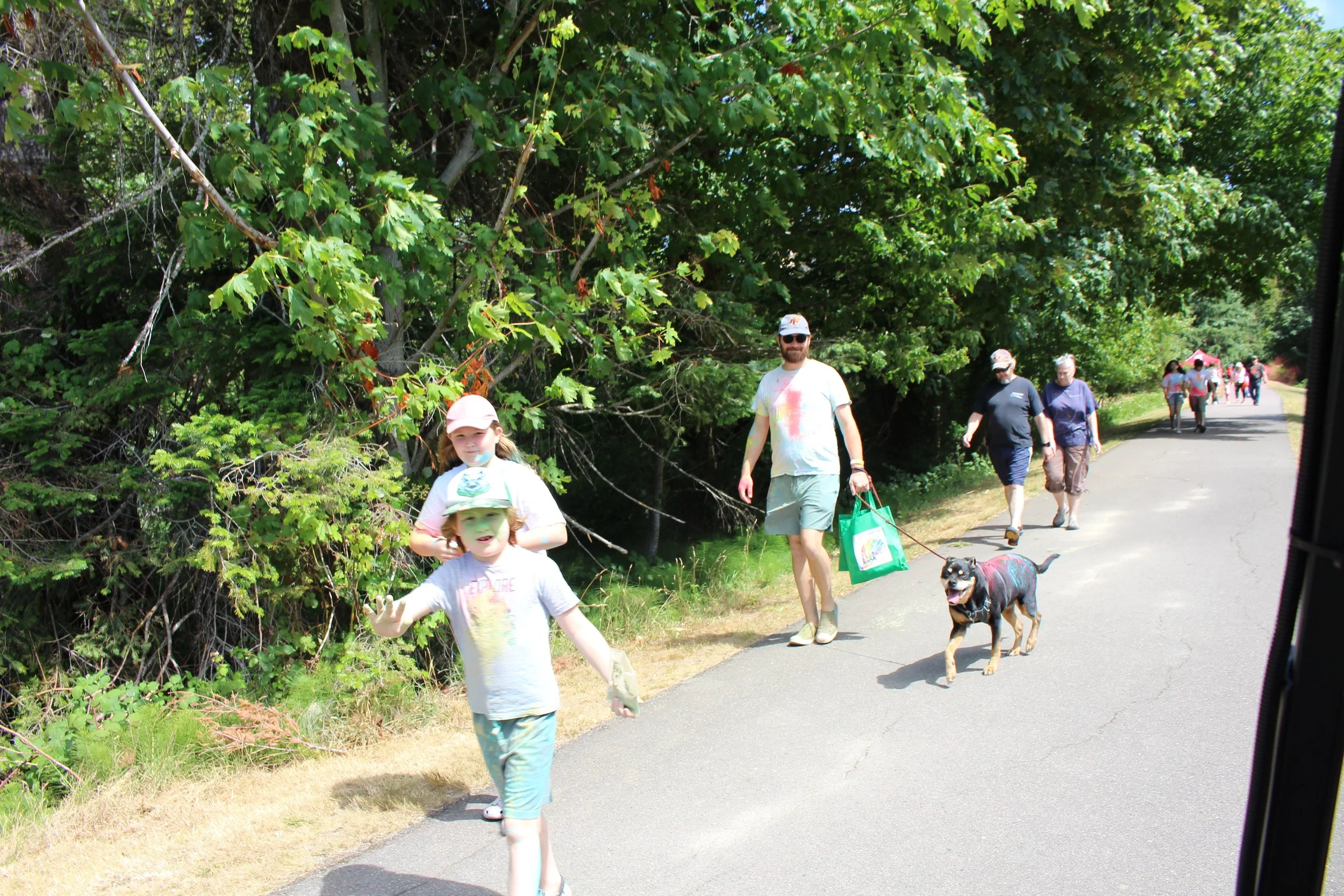 People walking along a paved trail near trees and greenery on a sunny day, including children and adults, one adult with a dog.