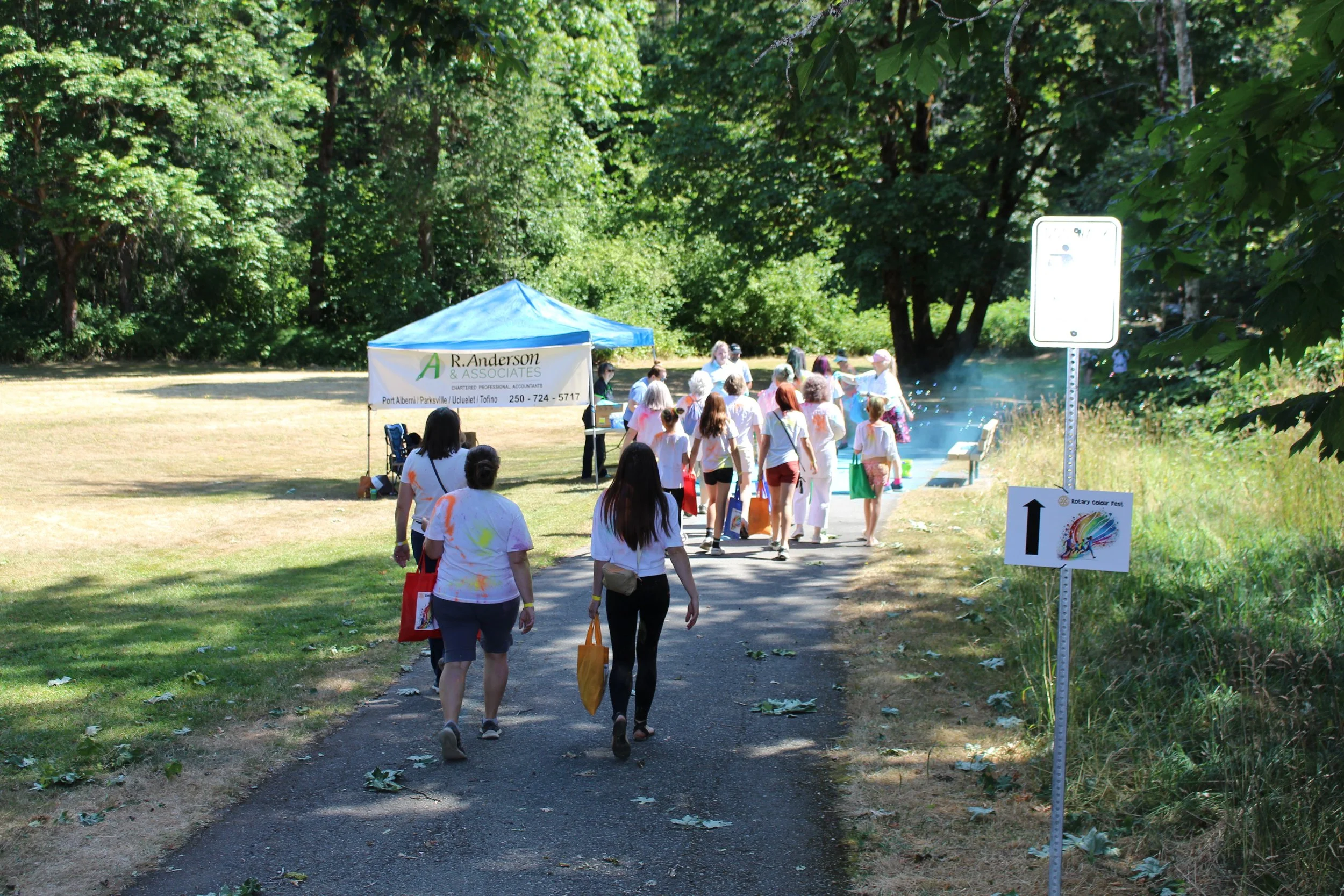 People walking on a paved pathway to an outdoor event station set up under a blue canopy tent, surrounded by green trees and grass, with signage and a small trail of scattered leaves.