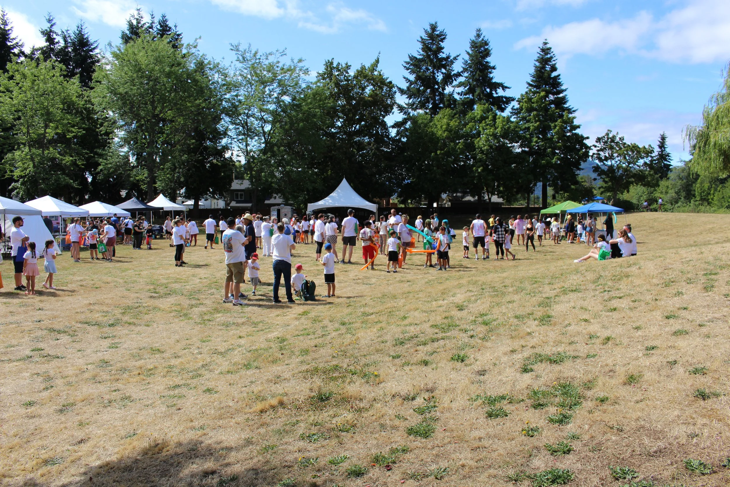 A park filled with families and children at an outdoor event, with tents and trees in the background on a sunny day.