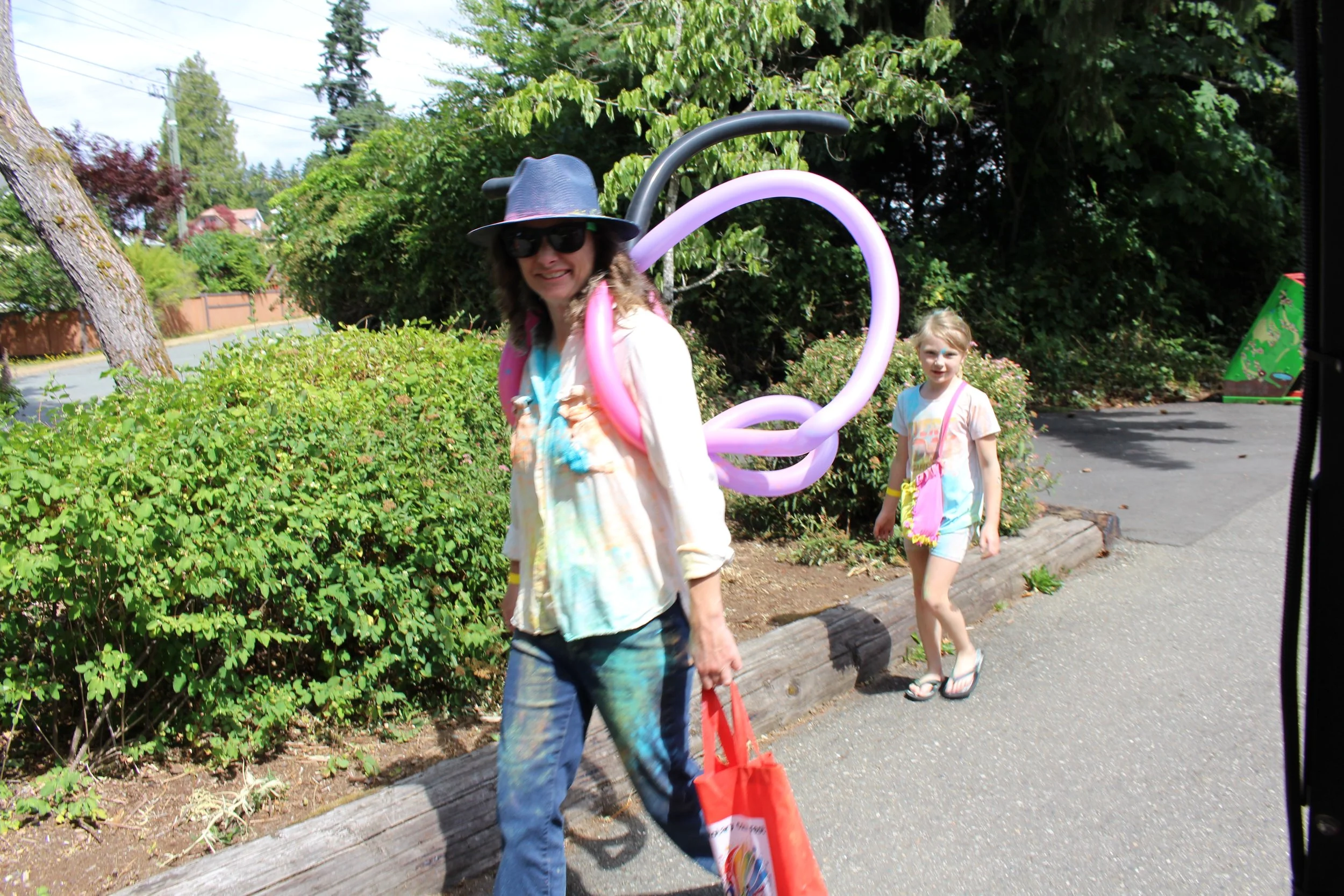 A woman and a young girl walking outside on a sidewalk, with the woman dressed casually in jeans, a colorful shirt, a hat, and sunglasses, carrying a red bag, and the girl wearing a T-shirt and shorts, carrying a pink bag, during daytime. There is a 